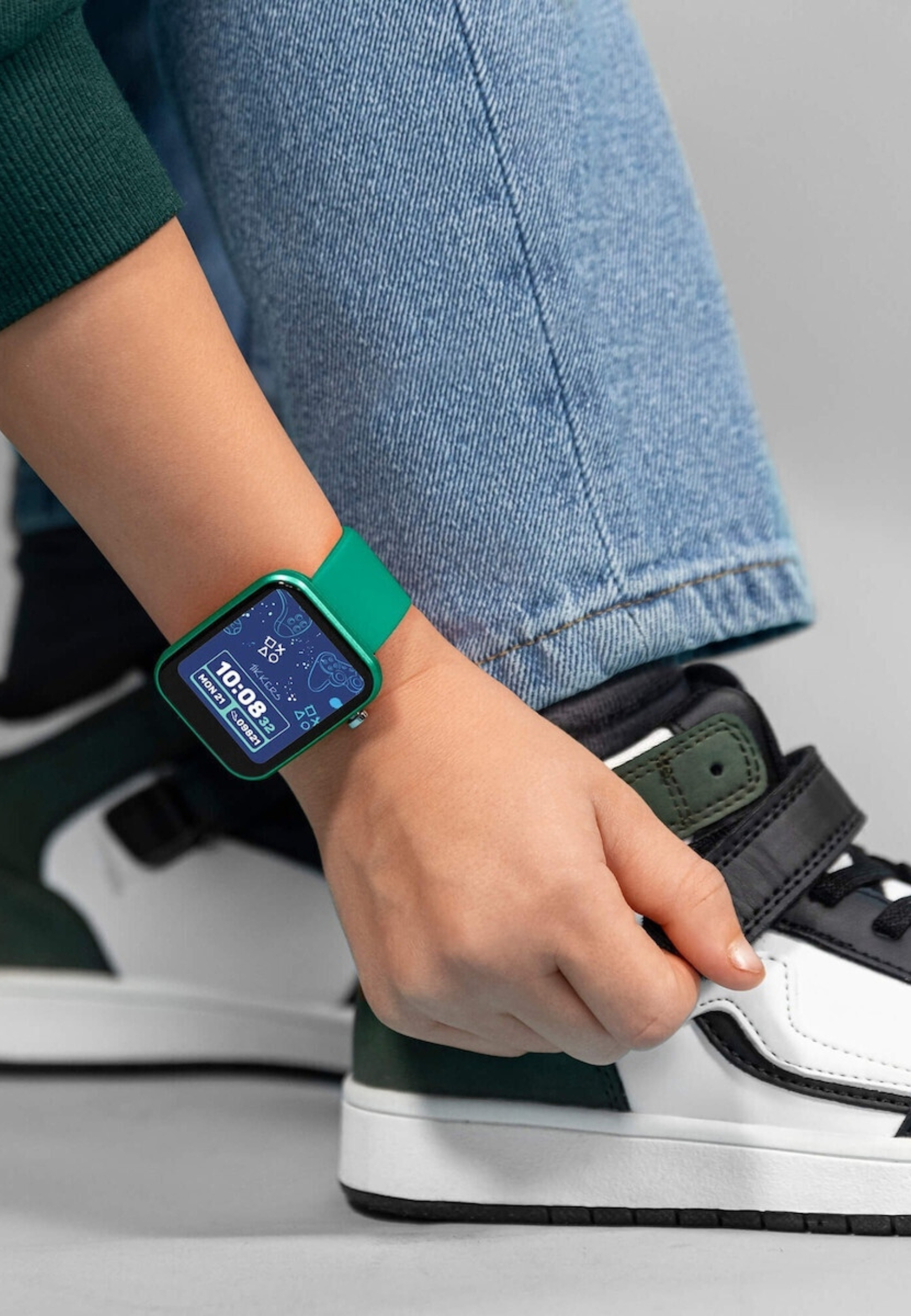 Ein Kinderarm mit einer grünen Smartwatch ist zu sehen. Das Kind trägt Jeans und schwarz-weiße Turnschuhe.