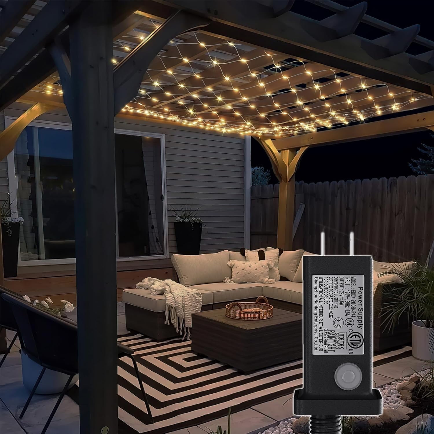 Eine Terrasse mit einer Pergola mit Lichterketten. Davor befinden sich ein Sofa, ein Couchtisch und ein Teppich. Ein Netzteil ist im Vordergrund.