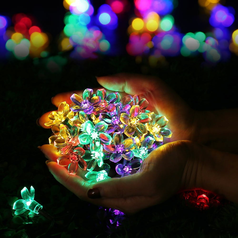 Hände umfassen leuchtende, bunte Blumenlichter. Der Hintergrund zeigt unscharfe Lichter. Ein einzelnes Blumenlicht ist im Vordergrund.