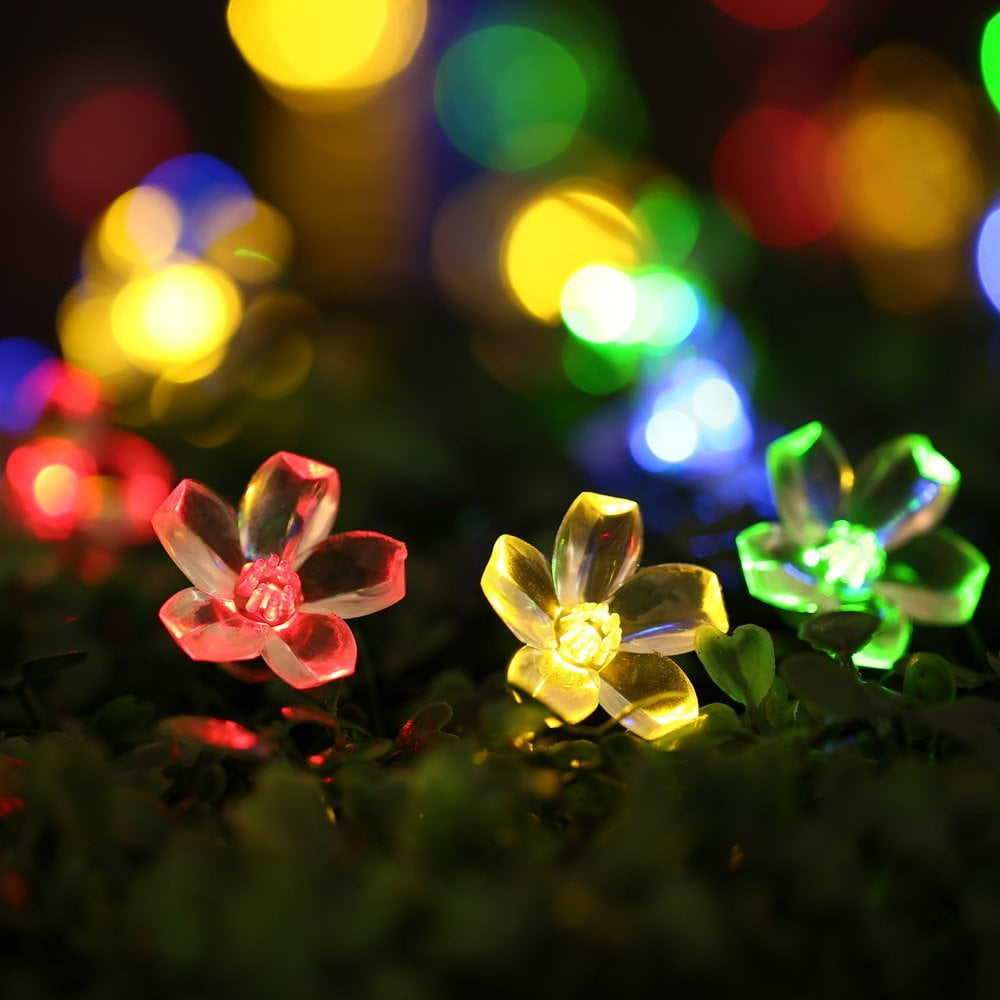 Nahaufnahme von blütenförmigen LED-Leuchten in Rot, Gelb und Grün auf grünem Laub. Verschwommene Bokeh-Lichter im Hintergrund.