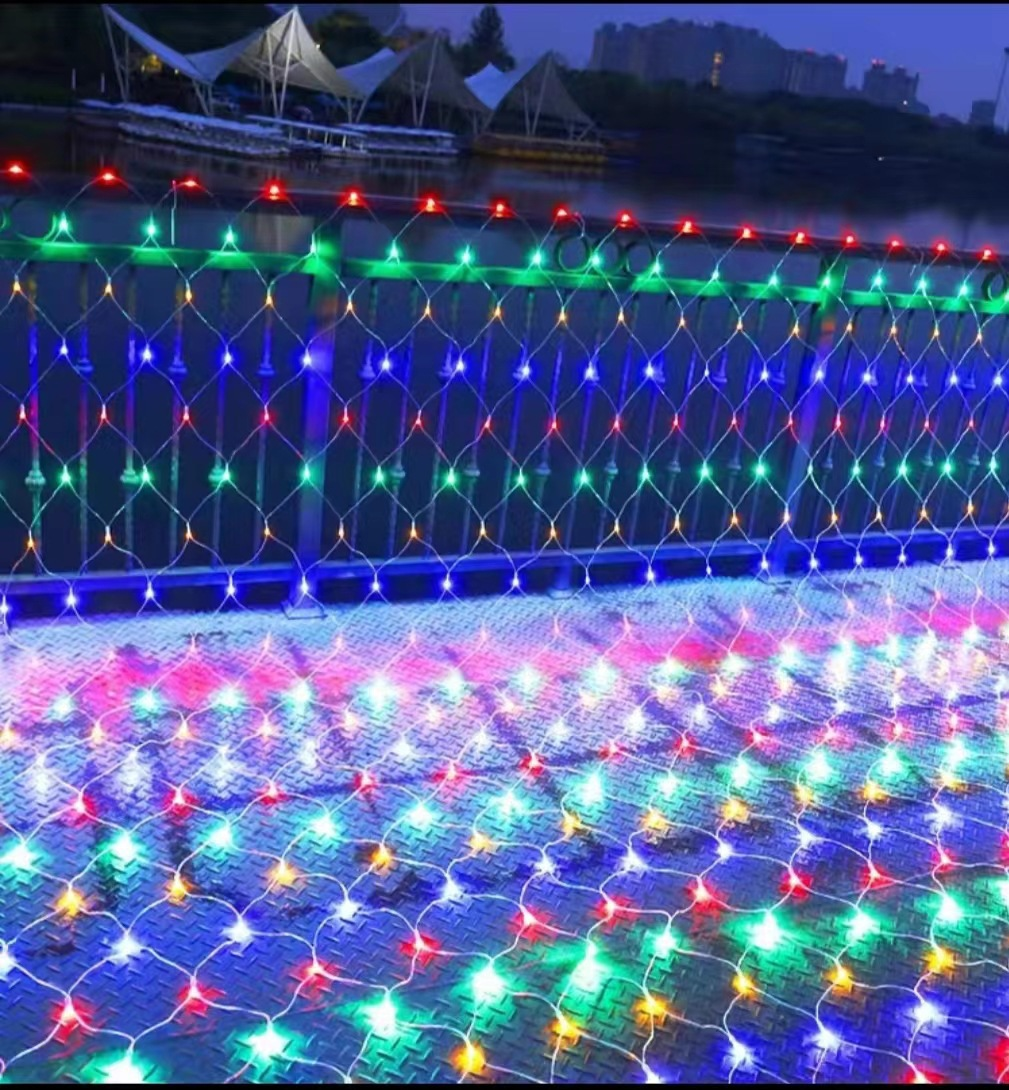 Eine Brücke mit bunten Netzlichtern geschmückt. Im Hintergrund sind Gebäude und Wasser. Die Lichter spiegeln sich am Boden.