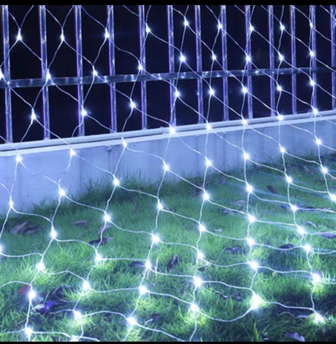 Ein Netz aus weißen Lichtern ist über Gras drapiert. Im Hintergrund ein Metallzaun. Die Lichter sind an und beleuchten die Szene.