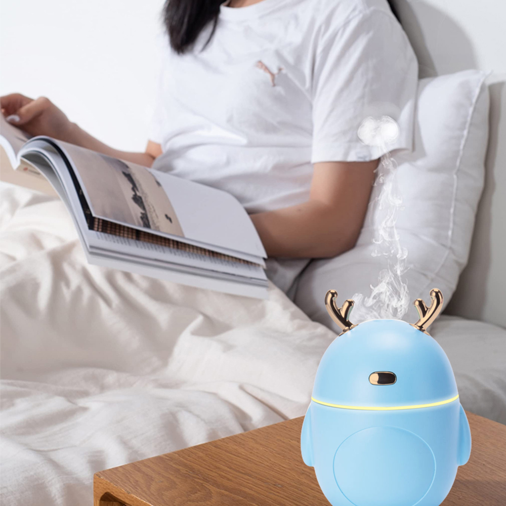Eine Person liest im Bett ein Buch. Ein blauer Luftbefeuchter mit goldenen Geweihen steht auf einem Holztisch und gibt Dampf ab. Der Hintergrund ist weiß.