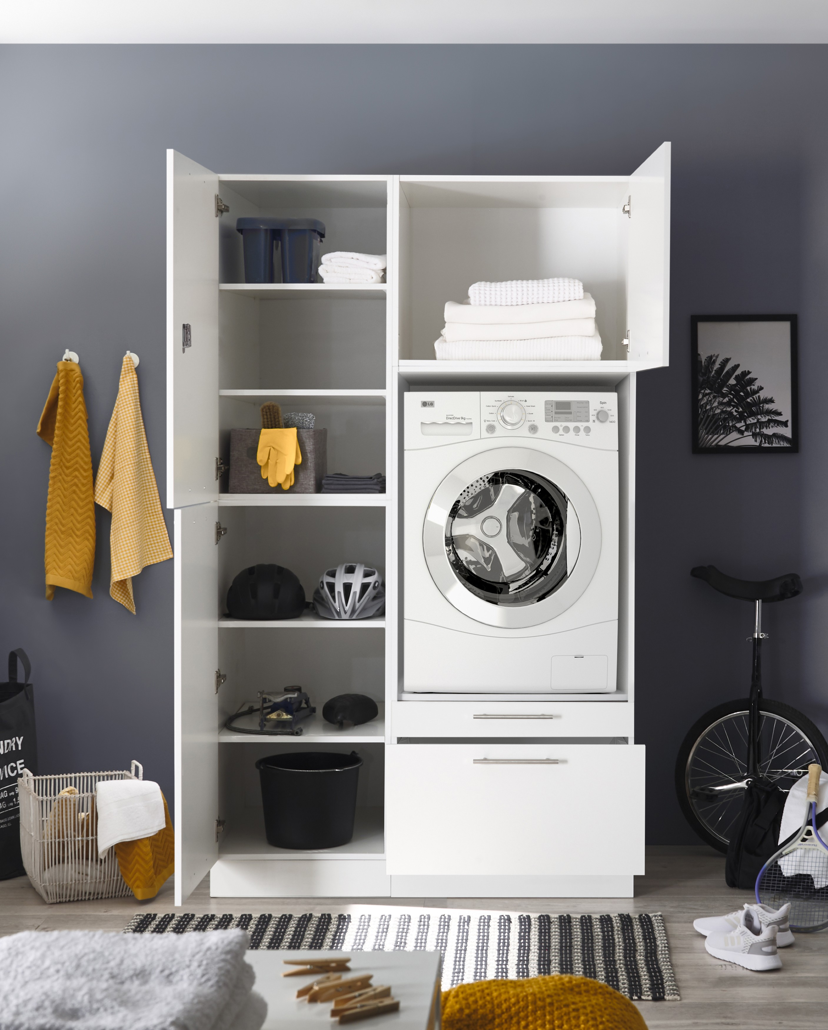 Ein weißer Wäscheschrank mit Waschmaschine. Regale halten Handtücher, Helme und Reinigungsmittel. Ein Einrad steht an der Wand.