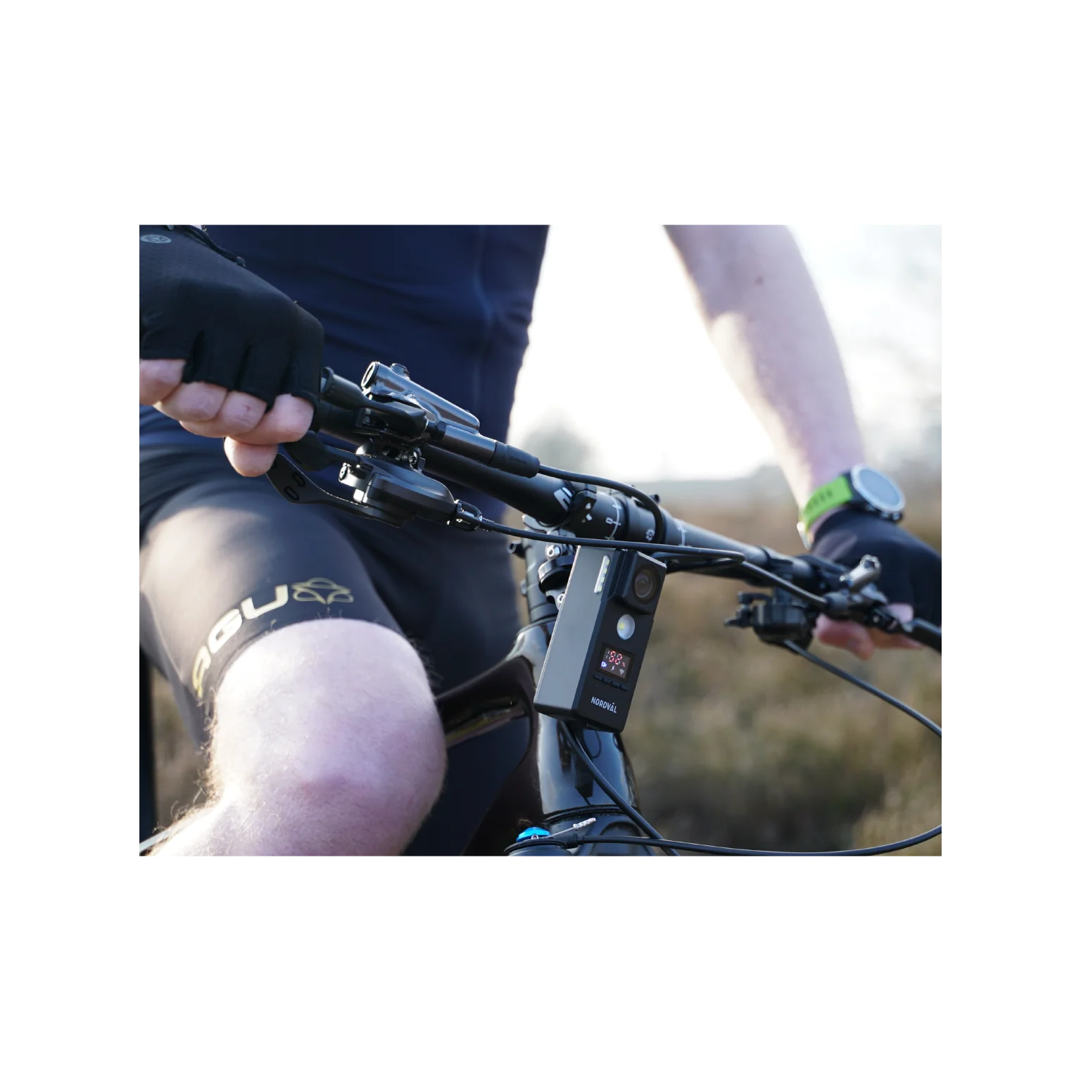 Cyclist on a bike, hands on handlebars. A watch, a bike light, and AGU shorts are visible. The scene is outdoors.