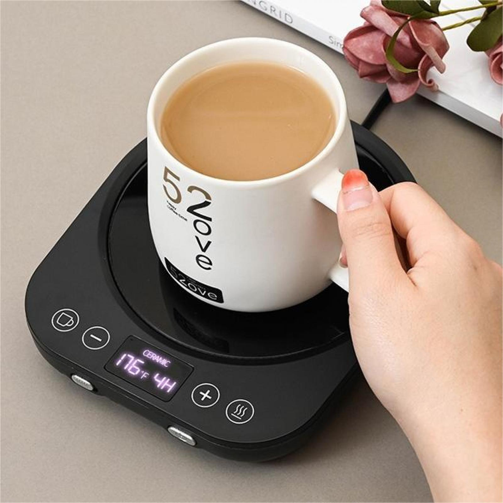 Eine weiße Tasse Kaffee auf einem schwarzen Wärmer, gehalten von einer Hand. Der Wärmer zeigt die Temperatur an. Blumen und ein Buch im Hintergrund.