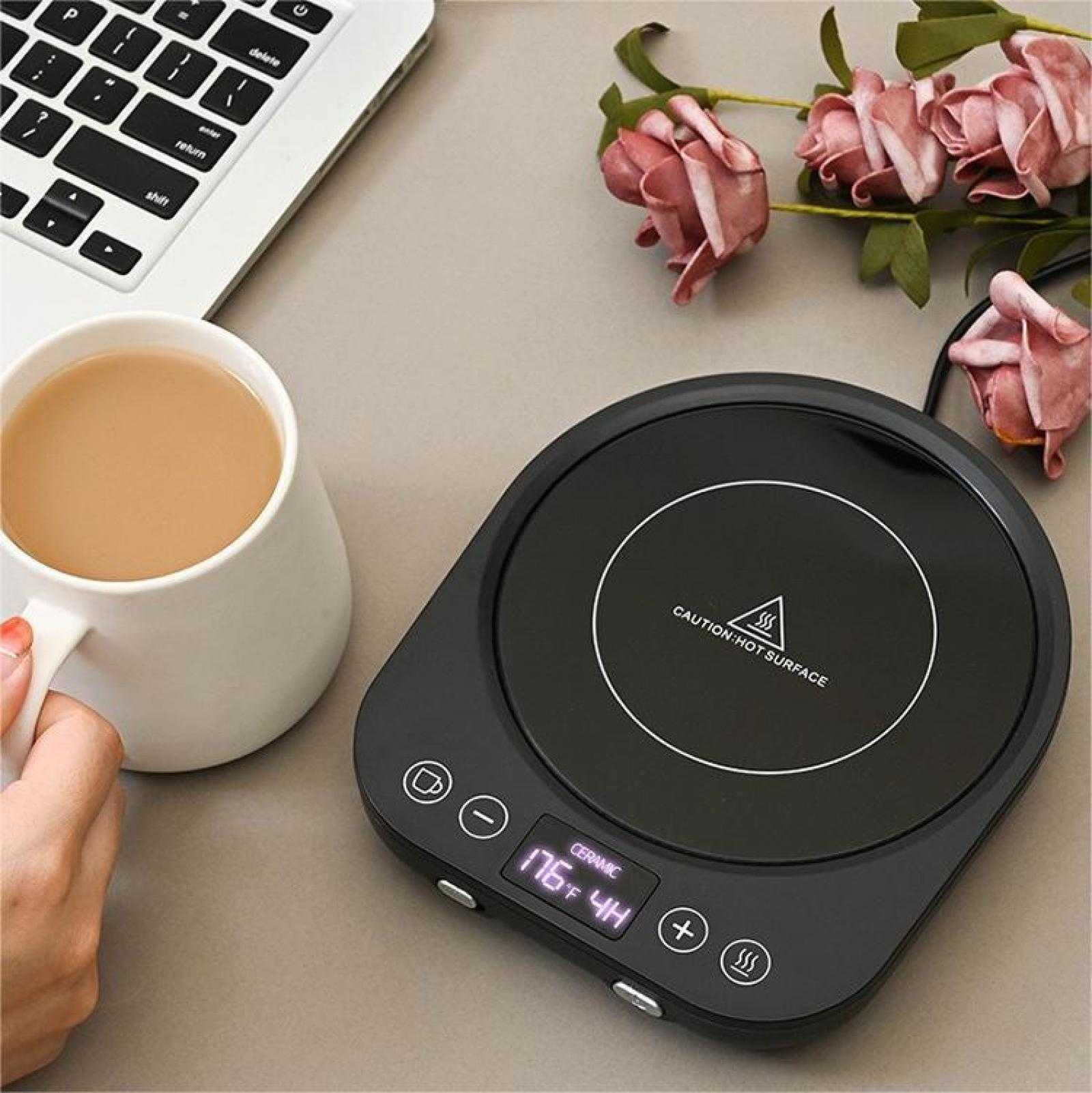 Eine weiße Tasse Kaffee, ein schwarzer Tassenwärmer mit Digitalanzeige, ein Laptop und rosa Blumen.