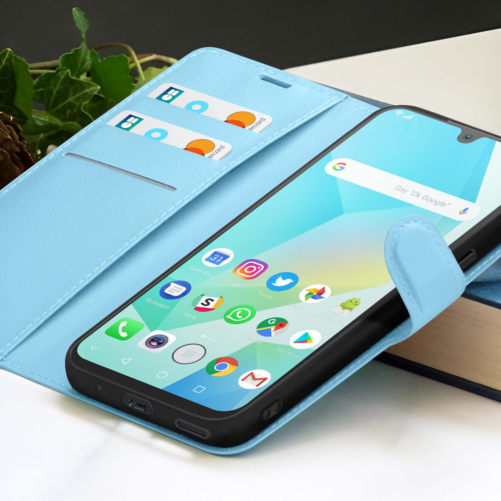Een smartphone in een lichtblauwe hoes met creditcards. Het scherm toont app-pictogrammen. Achtergrond omvat een boek en gebladerte.