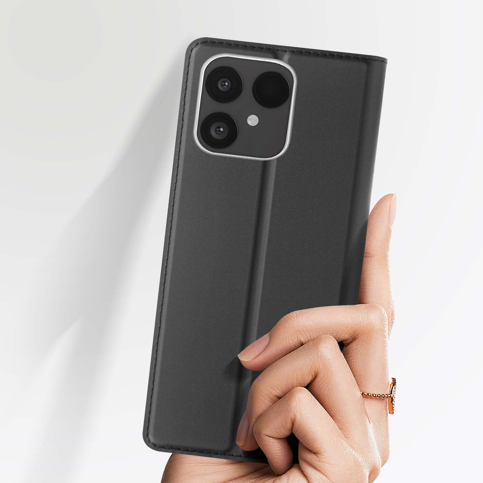 Una mano tiene una custodia nera per telefono con un modulo fotocamera. Un anello d'oro è sul dito. Lo sfondo è bianco.