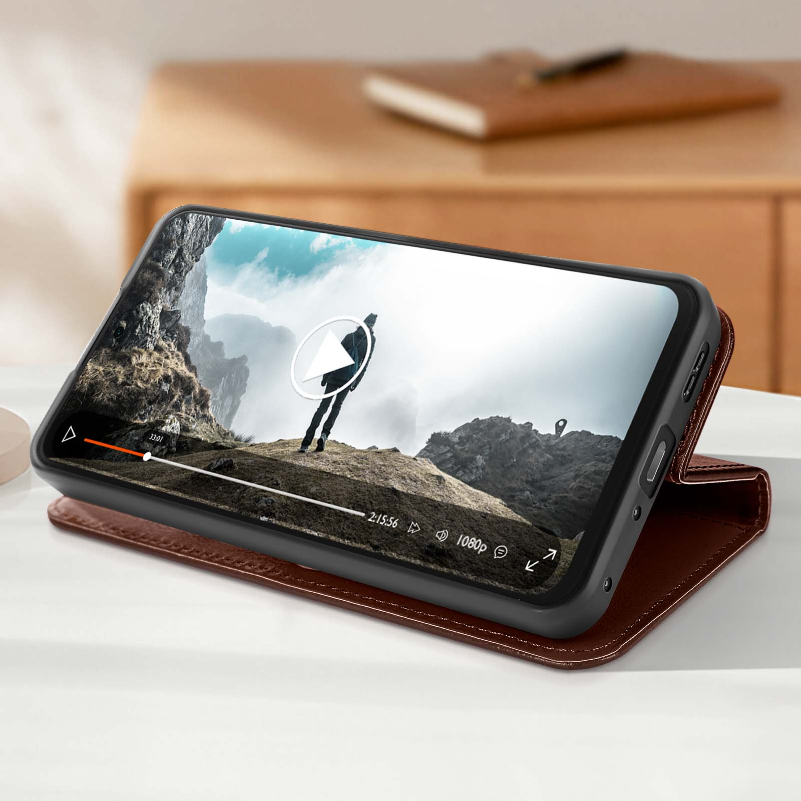 Un telefono in una custodia marrone mostra un video di una persona su una montagna. Un libro e una scrivania sullo sfondo.