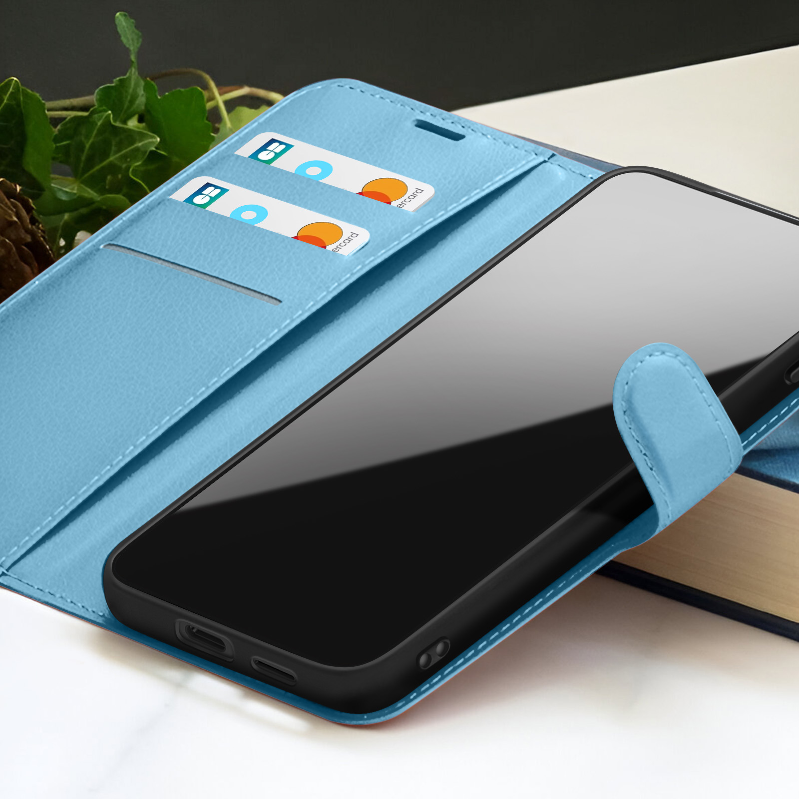 Un étui de téléphone bleu clair avec un téléphone noir à l'intérieur. Des cartes sont dans l'étui. Feuilles et un livre en arrière-plan.