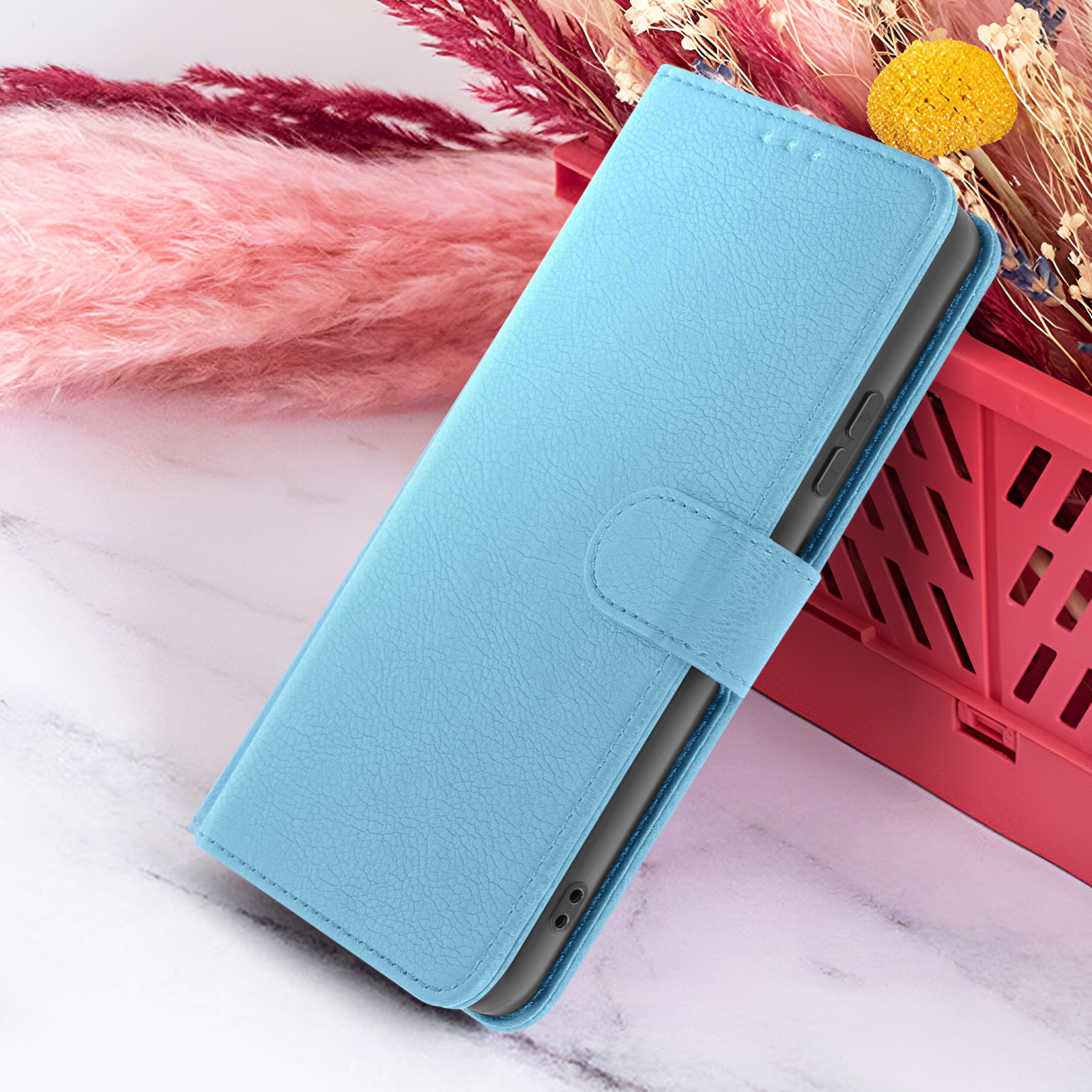Un étui de téléphone bleu clair avec fermeture magnétique est posé sur une surface en marbre. Des fleurs roses et bordeaux sont en arrière-plan.