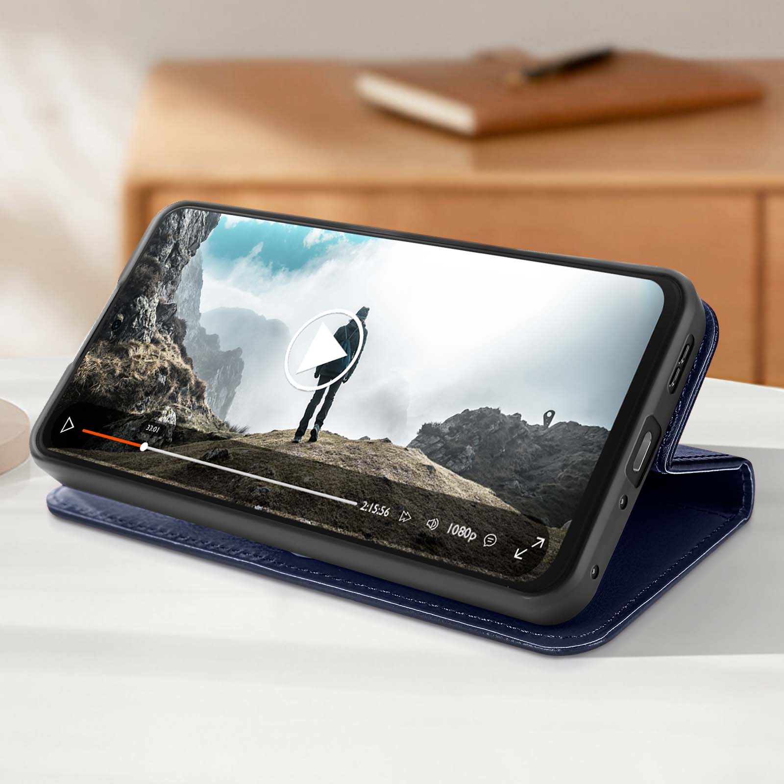 Uno smartphone in una custodia blu mostra un video di una persona che fa escursioni in montagna. Il telefono è su una superficie bianca.