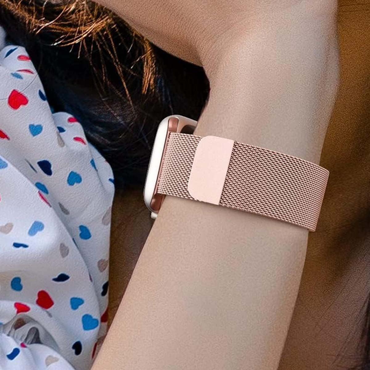 Uno smartwatch color oro rosa con un cinturino in rete abbinato è al polso di una persona. Una camicia fantasia è visibile sullo sfondo.