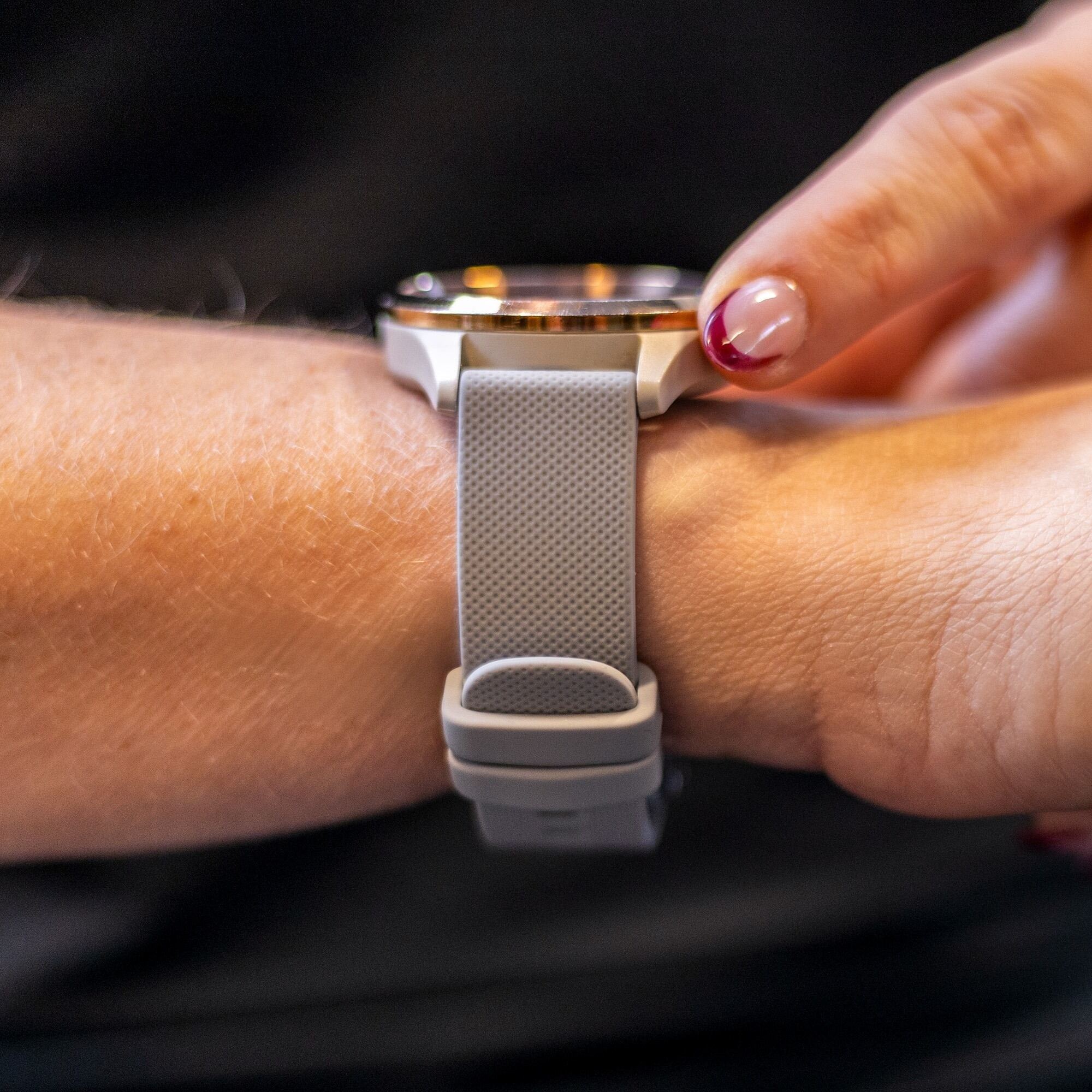 Arm einer Person mit grauer Uhr am Handgelenk. Ein Finger mit rotem Nagel berührt die Uhr.