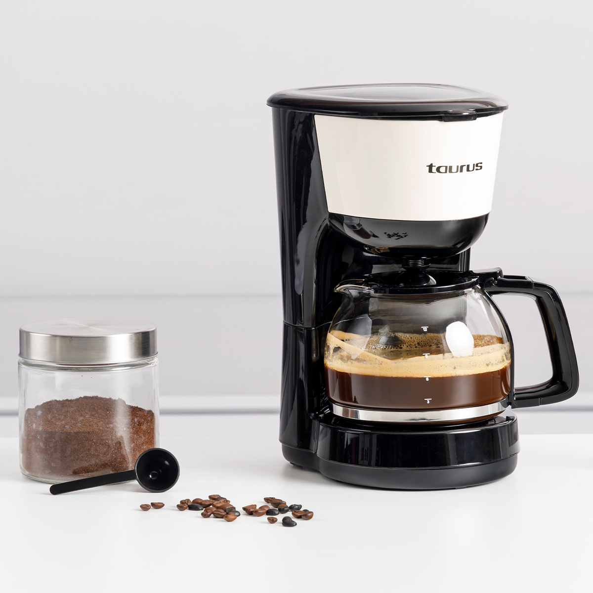 Una cafetera Taurus negra y blanca con una jarra de vidrio llena de café. Al lado, granos de café, una cuchara y un tarro de café.