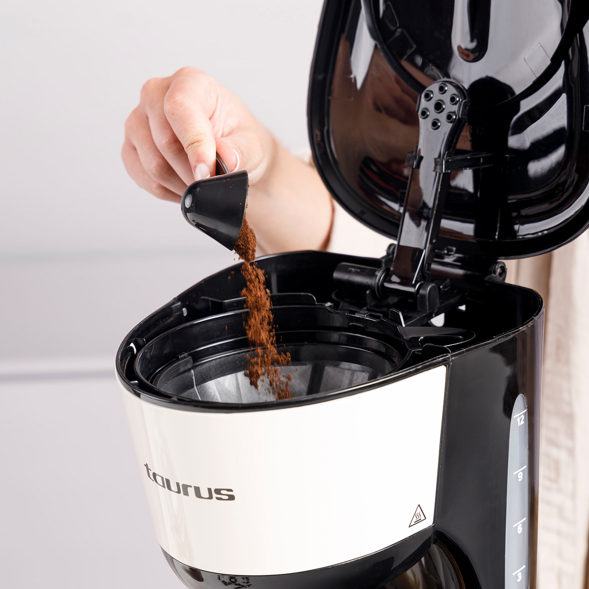 Una persona vierte café molido de una cuchara negra en una cafetera. La cafetera es blanca y negra.