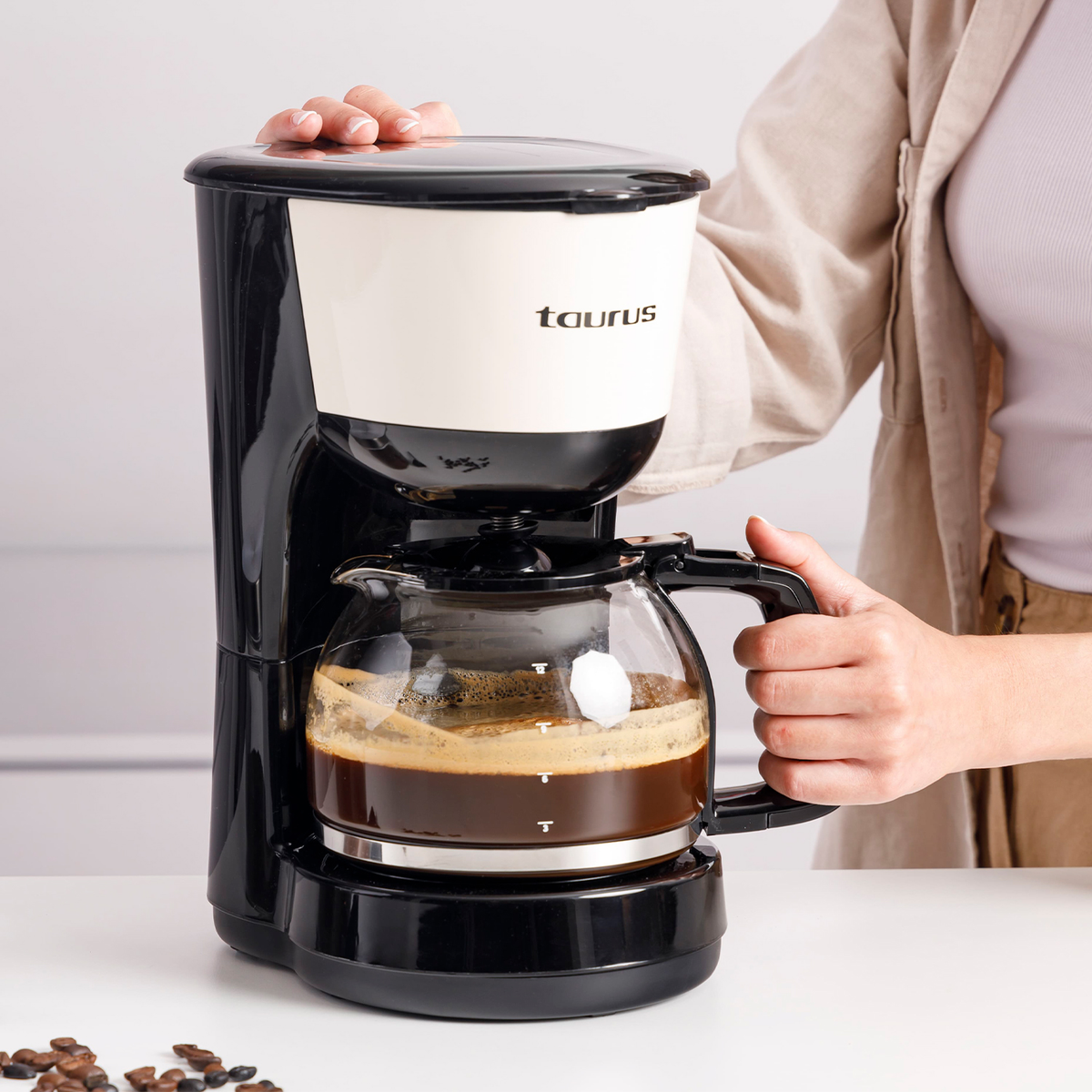 Una cafetera Taurus negra y blanca con una jarra de vidrio llena de café. Manos sostienen la jarra y la tapa.