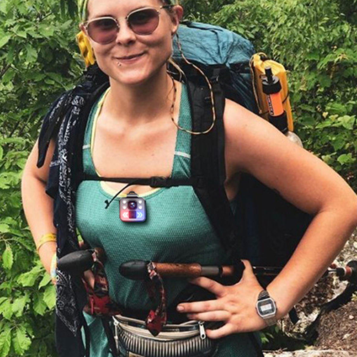 Frau mit Sonnenbrille, Rucksack und Kamera. Wandern in der Natur. Sie trägt ein grünes Tanktop und eine Uhr. Bäume im Hintergrund.
