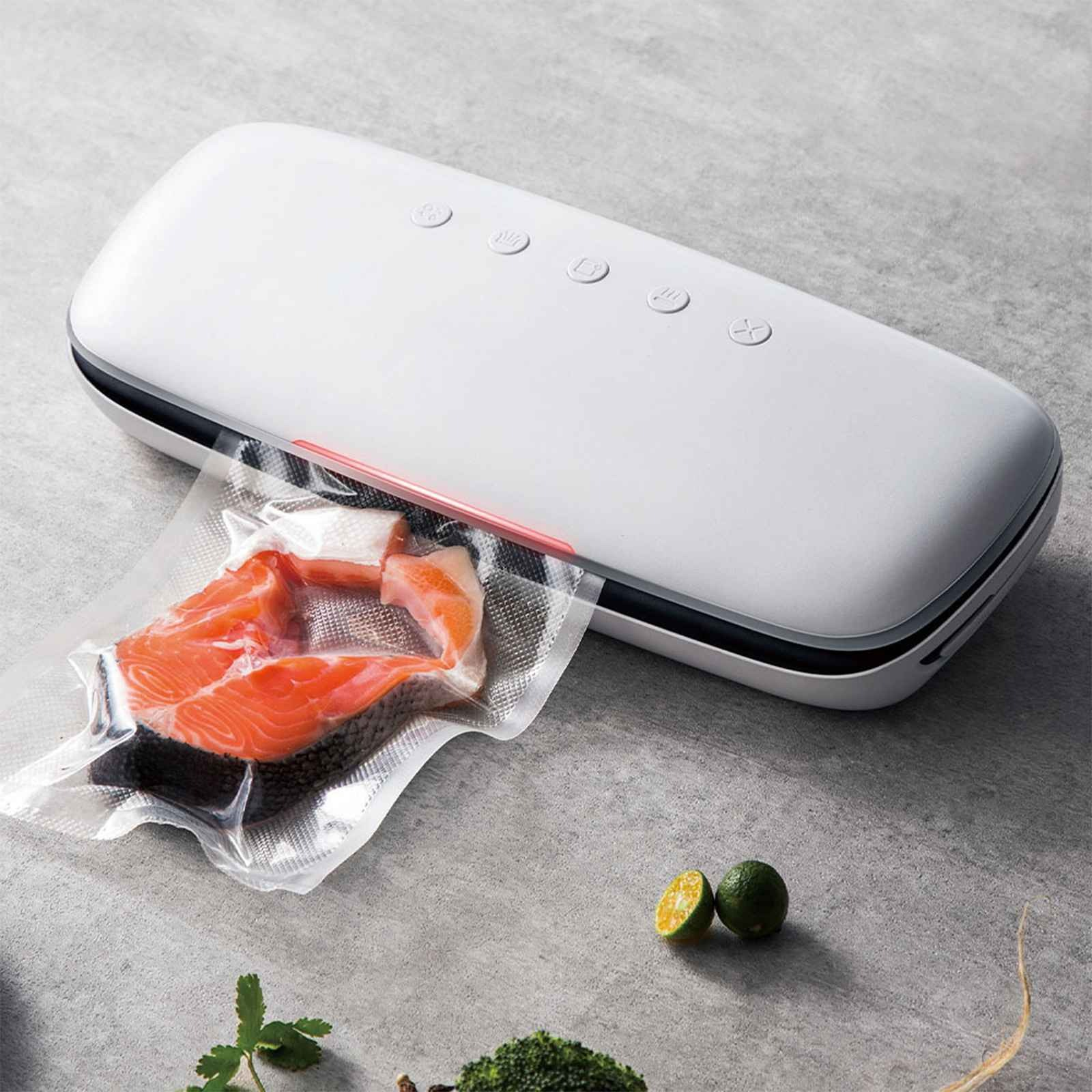 Ein weißes Vakuumiergerät mit Lachs in einer Tüte. Limetten, Kräuter und Brokkoli sind im Hintergrund. Die Oberfläche ist grau.