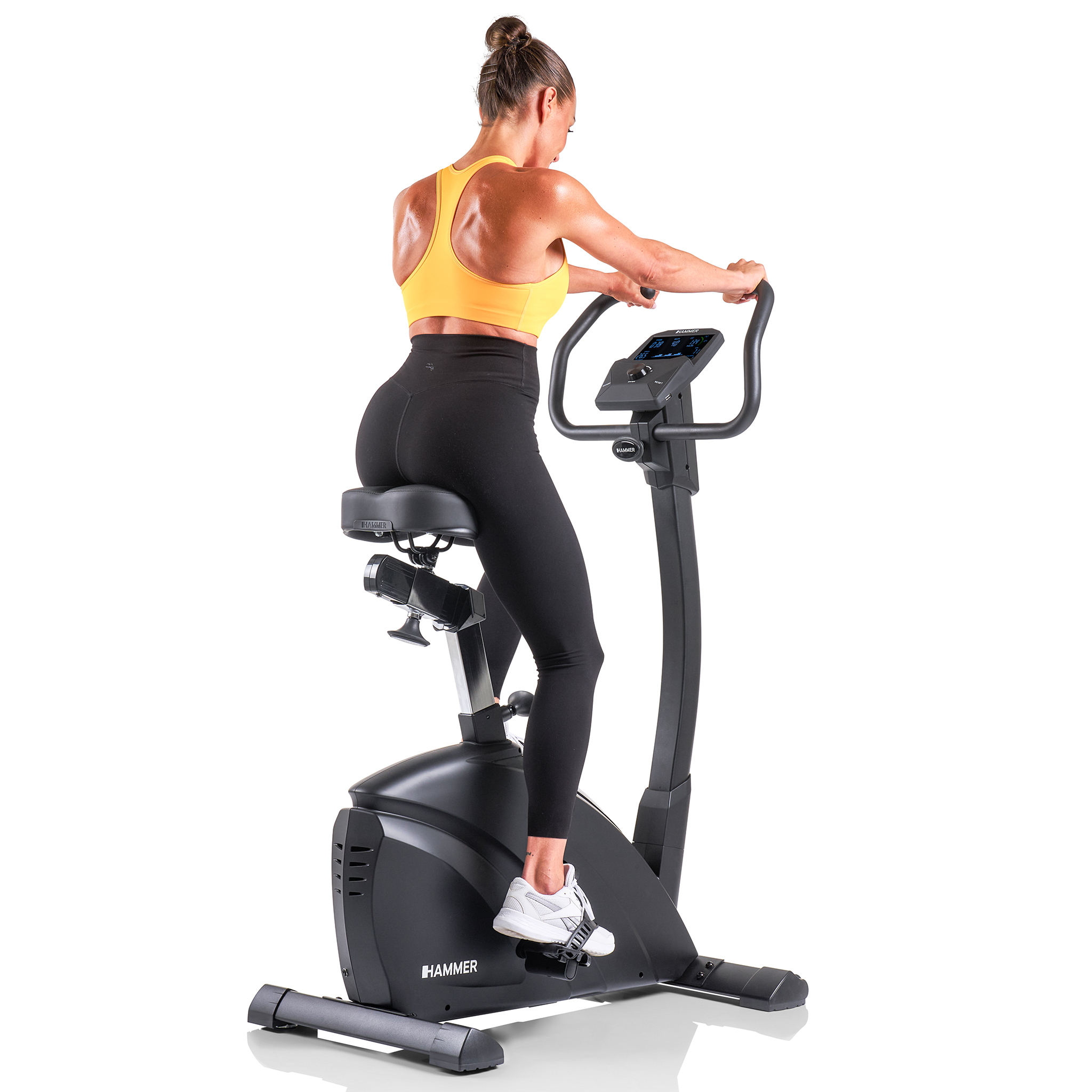 Frau auf dem Heimtrainer, gelbes Oberteil und schwarze Leggings. Rückansicht, Griff an den Griffen. Schwarzes Fahrrad, weißer Hintergrund.