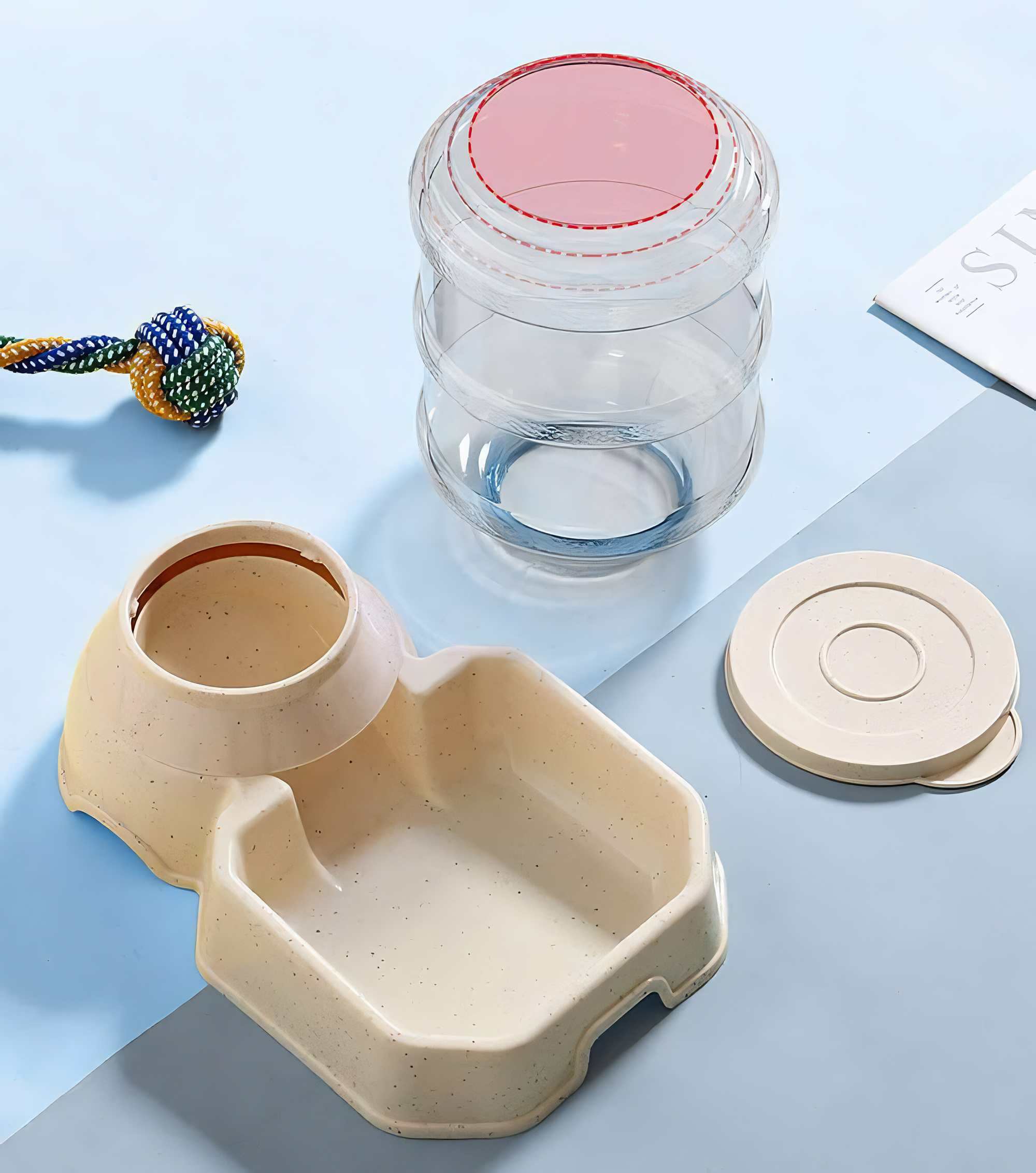 Ein Hunde-Wasserspender, eine Schüssel und ein Spielzeug auf einer hellblauen Oberfläche. Der Spender hat einen rosa Deckel. Das Spielzeug ist ein Seilknoten.