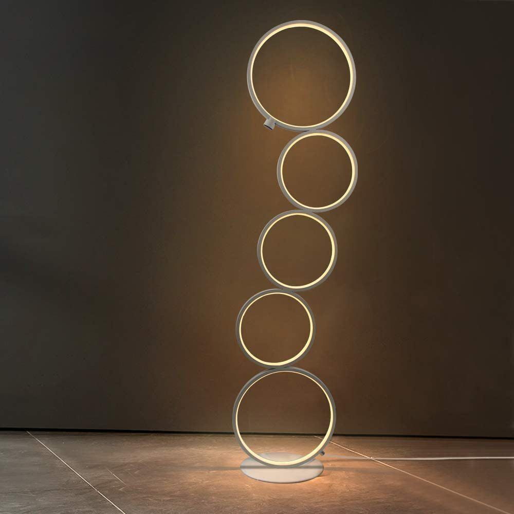 Eine moderne Stehlampe mit verbundenen, beleuchteten Ringen. Die Lampe hat einen weißen Sockel und eine dunkle Wand im Hintergrund.