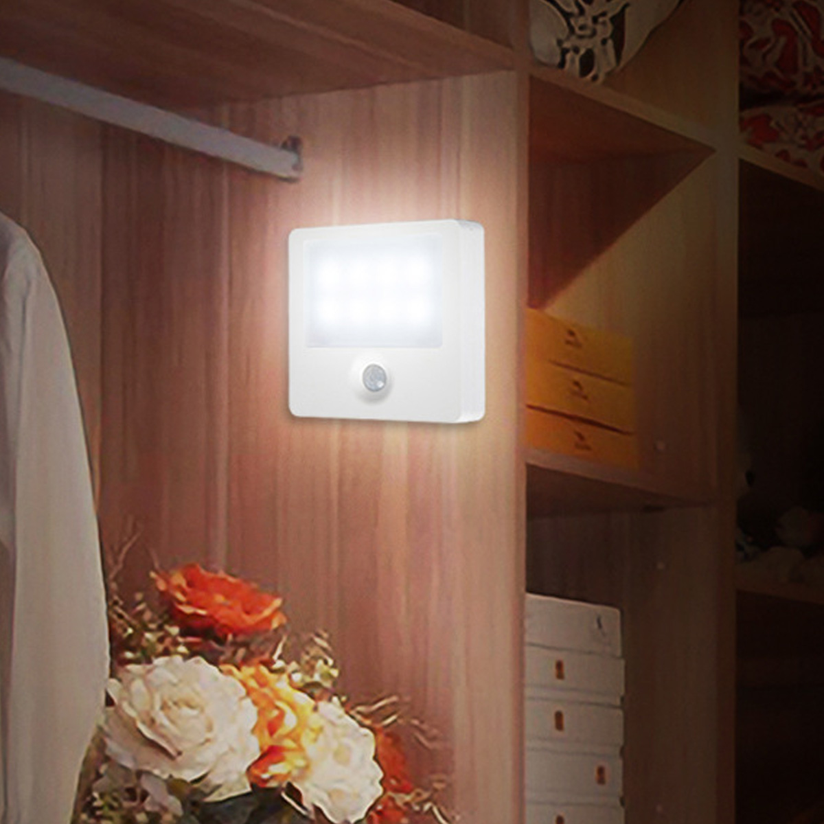 Ein weißes LED-Licht mit Bewegungssensor ist an einer Holzwand montiert. Ein Blumenstrauß steht darunter. Rechts sind Regale zu sehen.