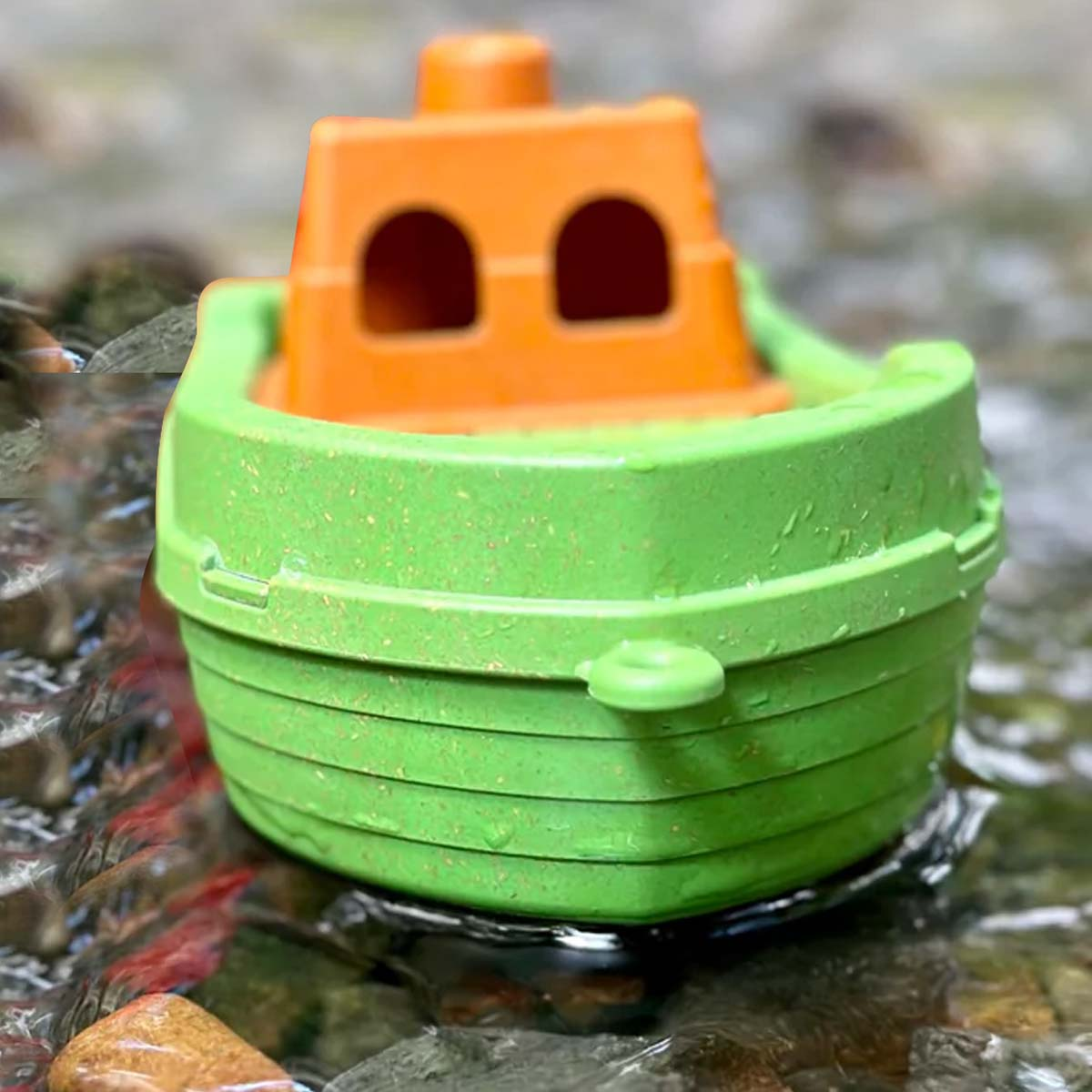 Ein grünes Spielzeugboot mit einer orangefarbenen Kabine schwimmt in einem Bach. Wasser fließt um das Boot, und es gibt Felsen im Wasser.