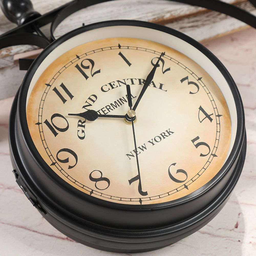 Eine schwarze, doppelseitige Uhr zeigt 'Grand Central Terminal New York' an. Zeiger stehen auf 1:06. Die Uhr steht auf einer weißen und rosa Oberfläche.