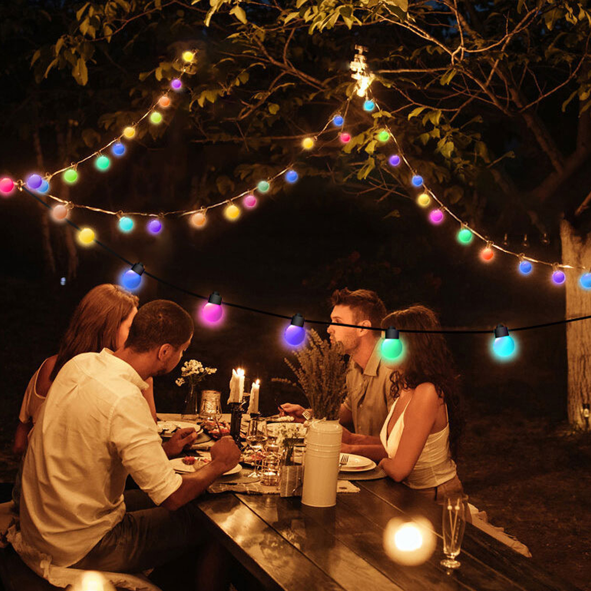 Menschen speisen im Freien unter bunten Lichterketten. Ein Tisch ist mit Essen und Kerzen gedeckt. Bäume und dunkler Himmel im Hintergrund.