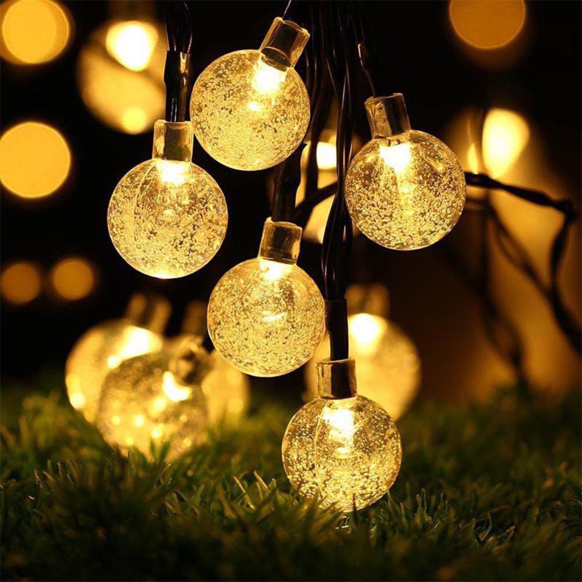 Nahaufnahme von leuchtenden, kugelförmigen Lichterketten. Die Lichter sind an schwarzen Drähten, mit Gras im Vordergrund und Bokeh-Lichtern im Hintergrund.