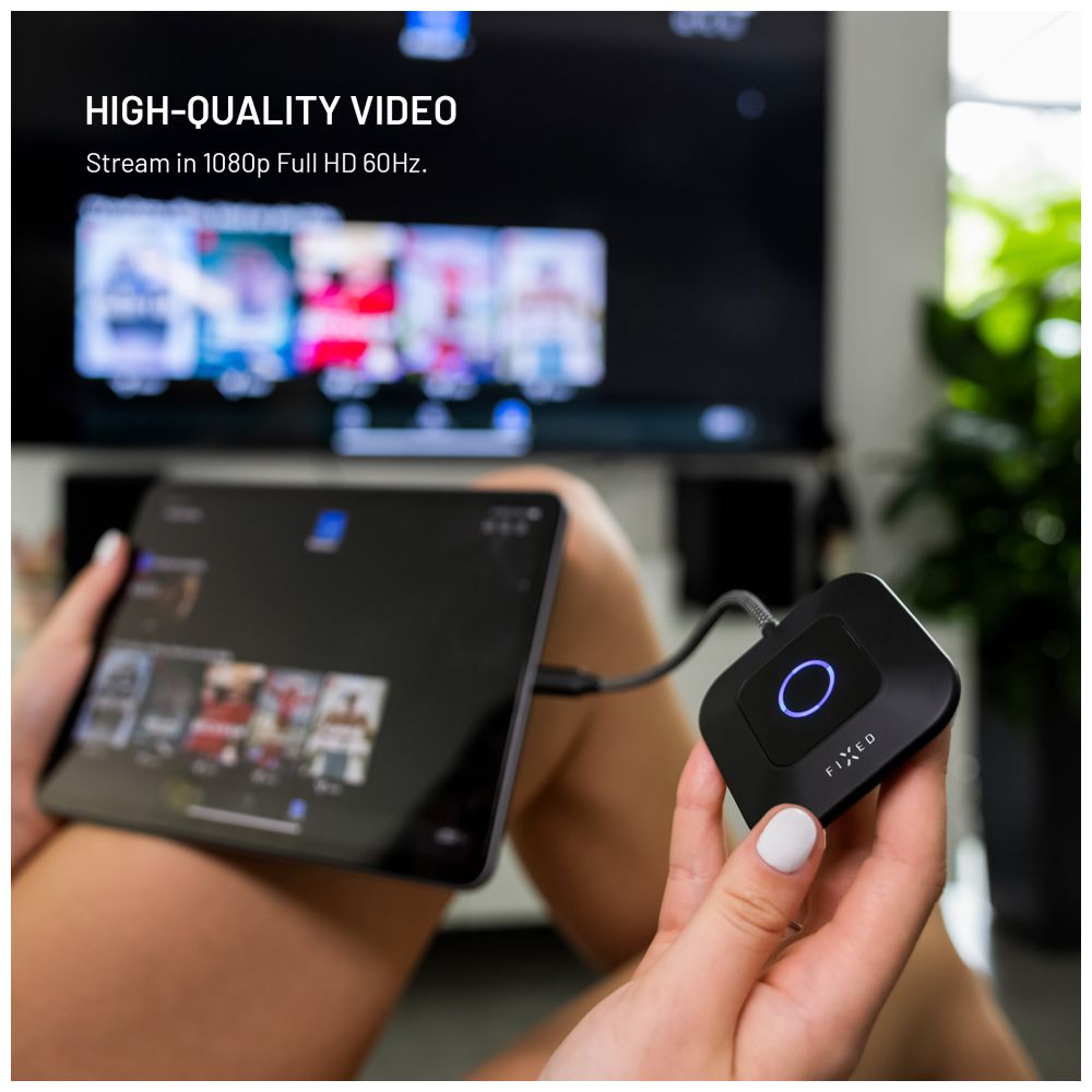 Eine Person hält ein Tablet, das mit einem schwarzen Gerät mit blauem Licht verbunden ist. Der Hintergrund zeigt einen Fernseher und grüne Pflanzen.
