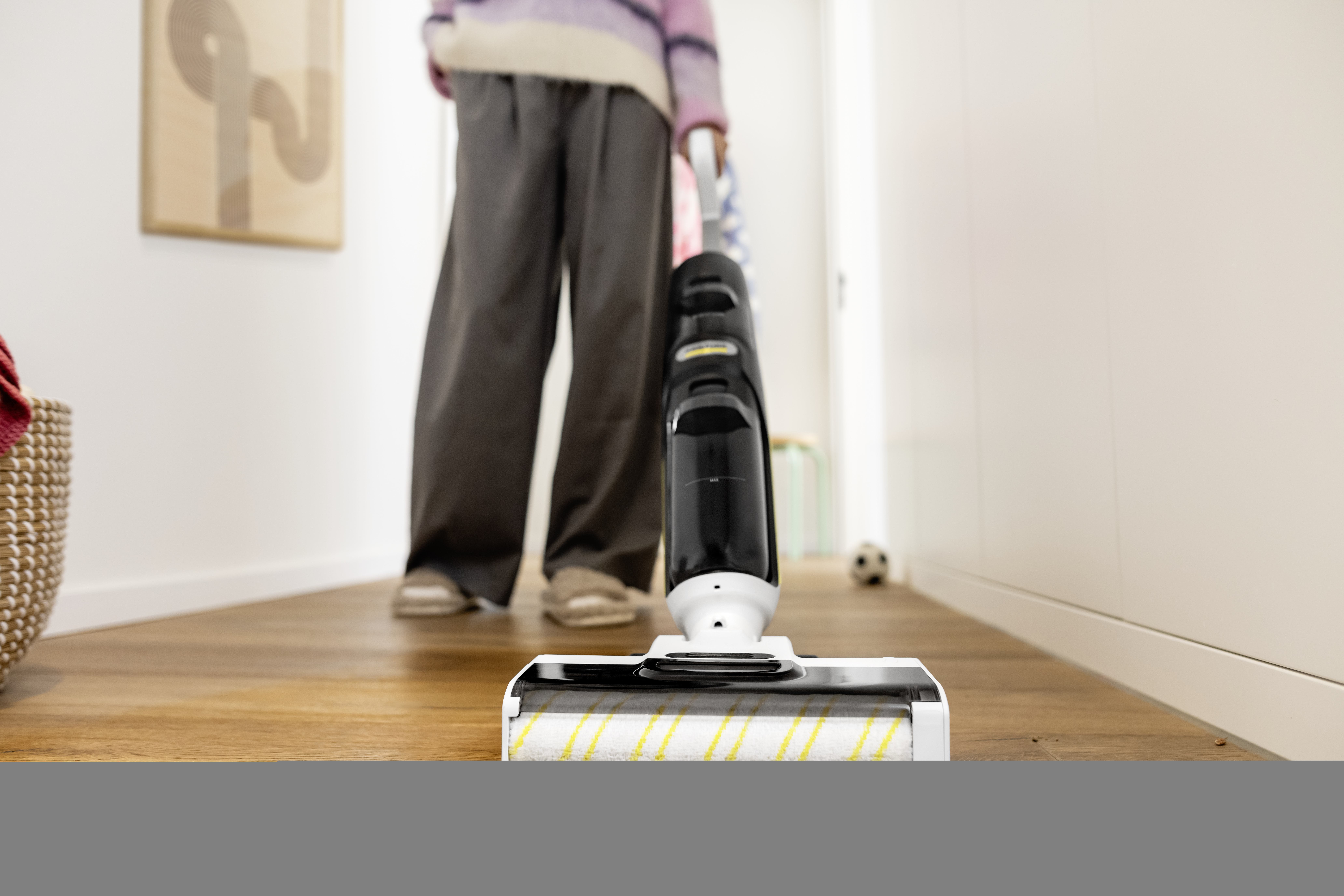 Person using a black and white vacuum cleaner on a wooden floor. A painting hangs on the wall. A ball is in the background.