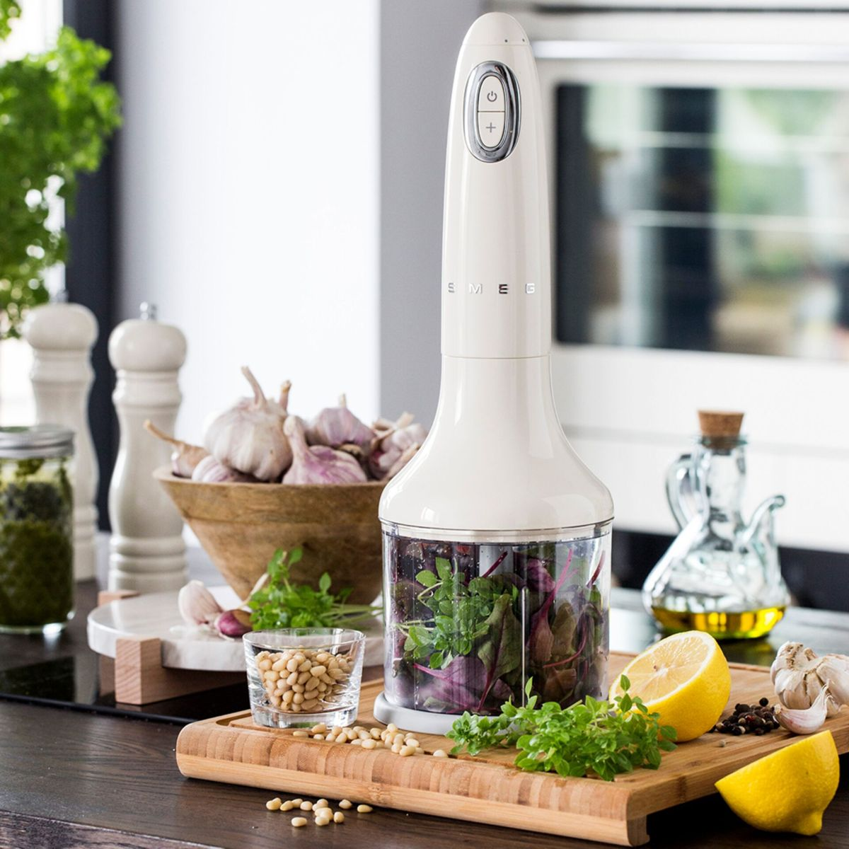 Kremowy blender ręczny obok składników takich jak czosnek, cytryna, zioła i orzeszki piniowe na drewnianej desce. Widoczny słoik pesto.