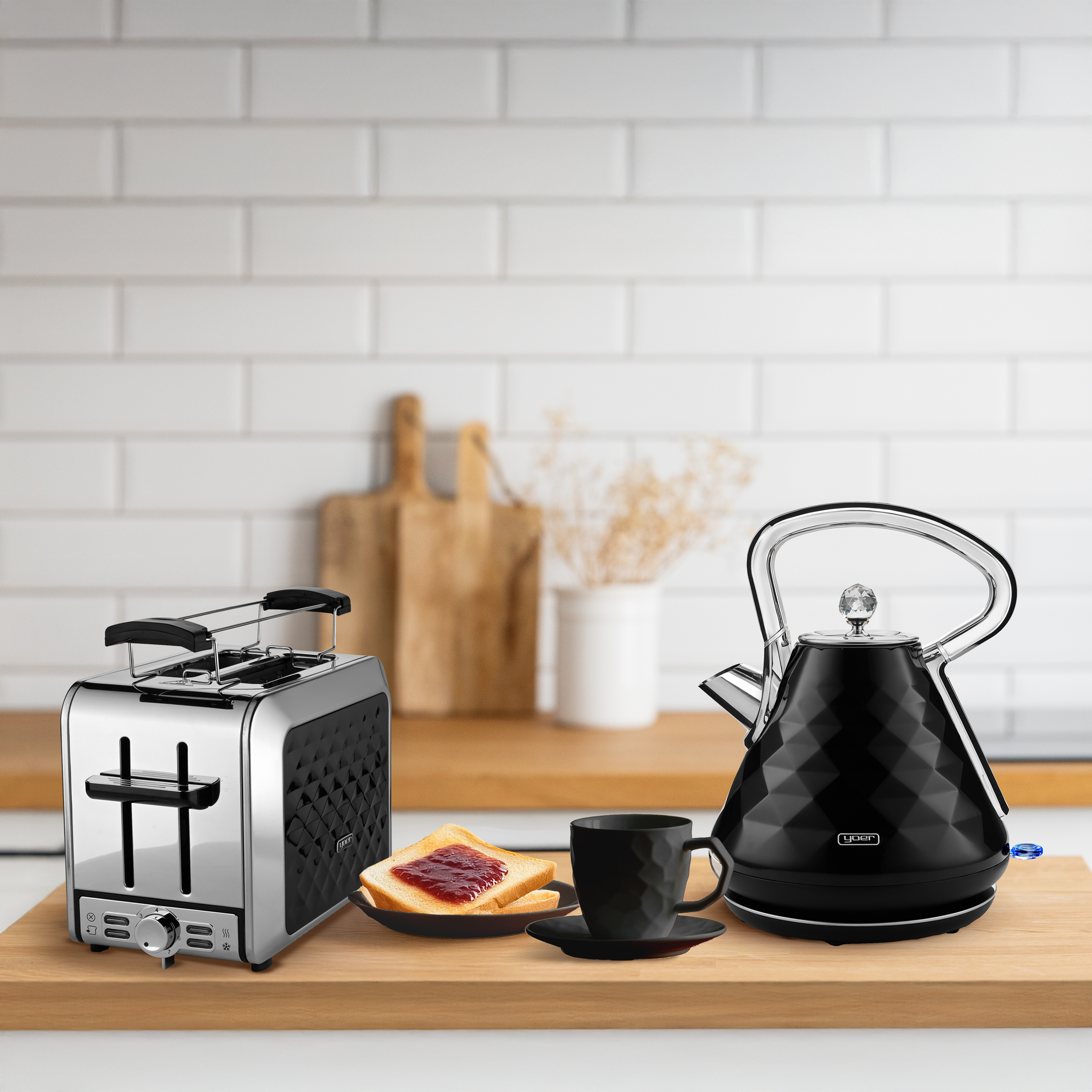 Chromowany toster, czarny czajnik, filiżanka ze spodkiem i tosty z dżemem na drewnianej desce.