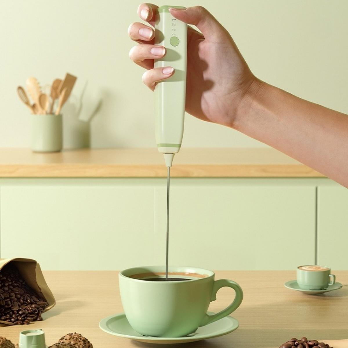 Eine Hand hält einen Milchaufschäumer über einer grünen Kaffeetasse. Kaffeebohnen, eine kleine Tasse und Küchenutensilien liegen auf dem Tisch.