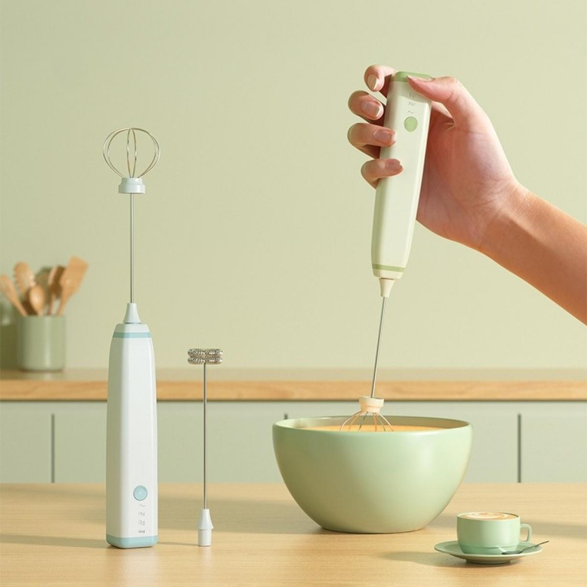 Eine Hand hält einen weißen und grünen Mixer über einer Schüssel mit Flüssigkeit. Zwei Mixer und eine Tasse Kaffee stehen auf einem Holztisch.
