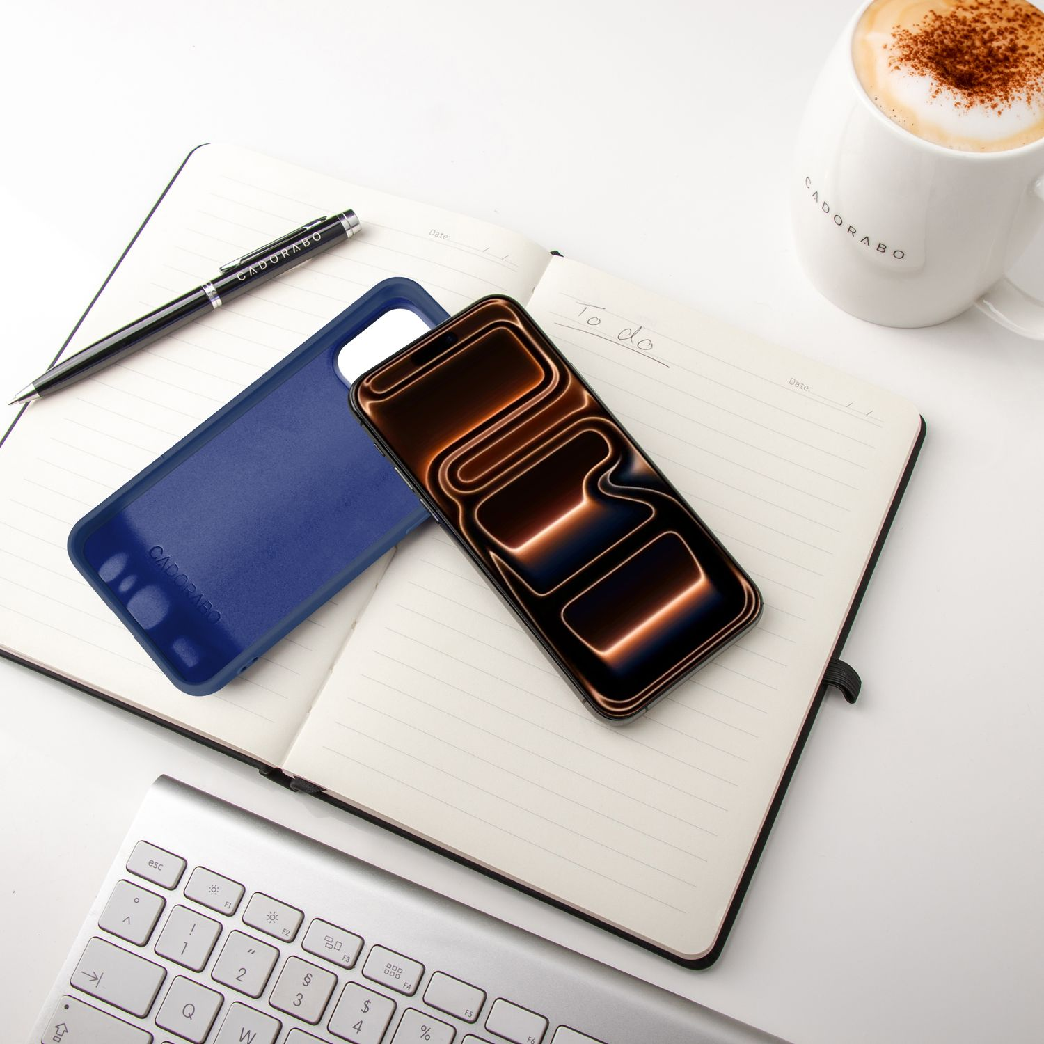 Notizbuch, Telefon, Stift, blaue Handyhülle und Kaffeetasse auf einem weißen Schreibtisch. Das Telefon zeigt ein Muster. Eine Tastatur ist sichtbar.