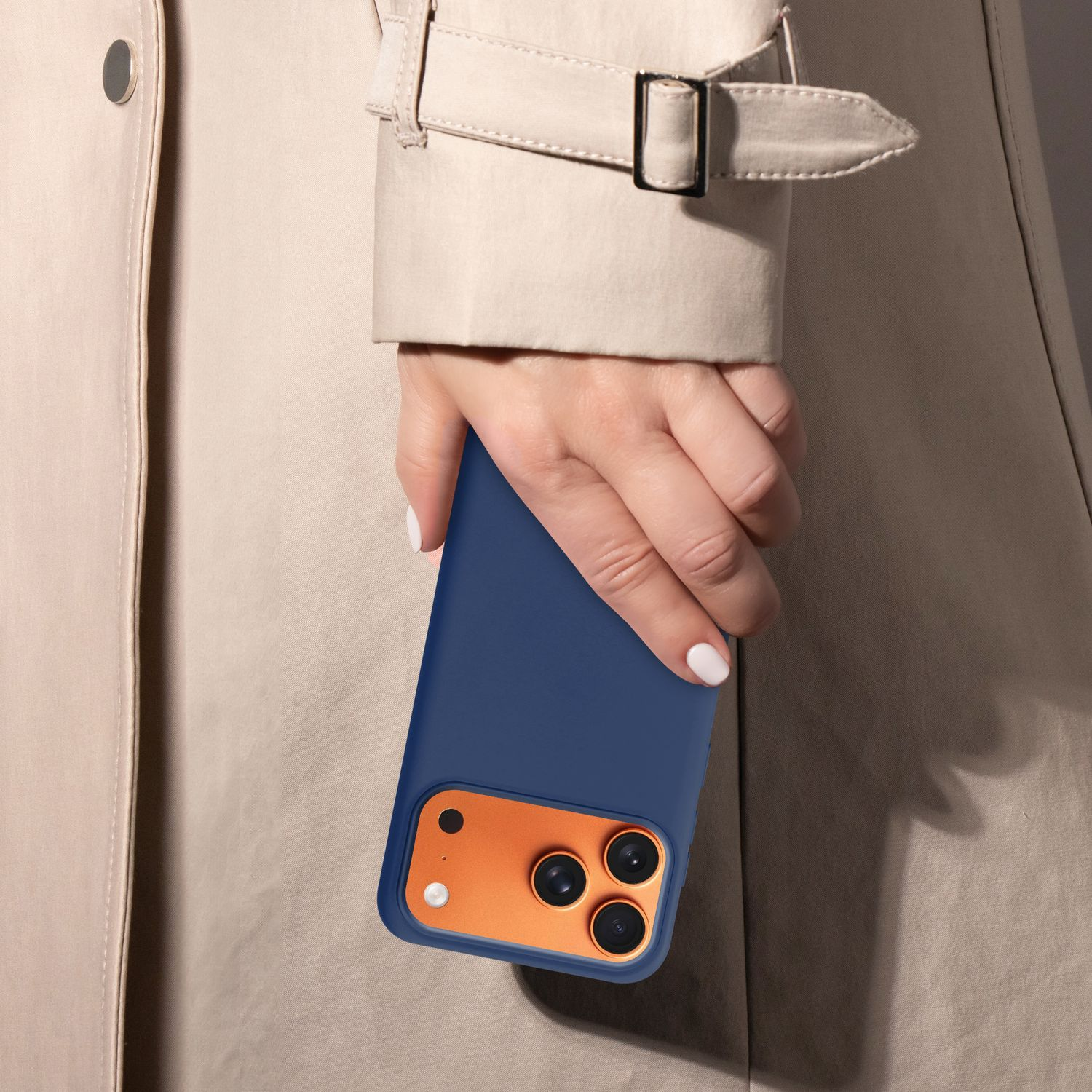 Eine Person hält ein blaues Telefon mit einem orangefarbenen Kameramodul. Die Person trägt einen beigefarbenen Trenchcoat. Weißer Nagellack ist sichtbar.