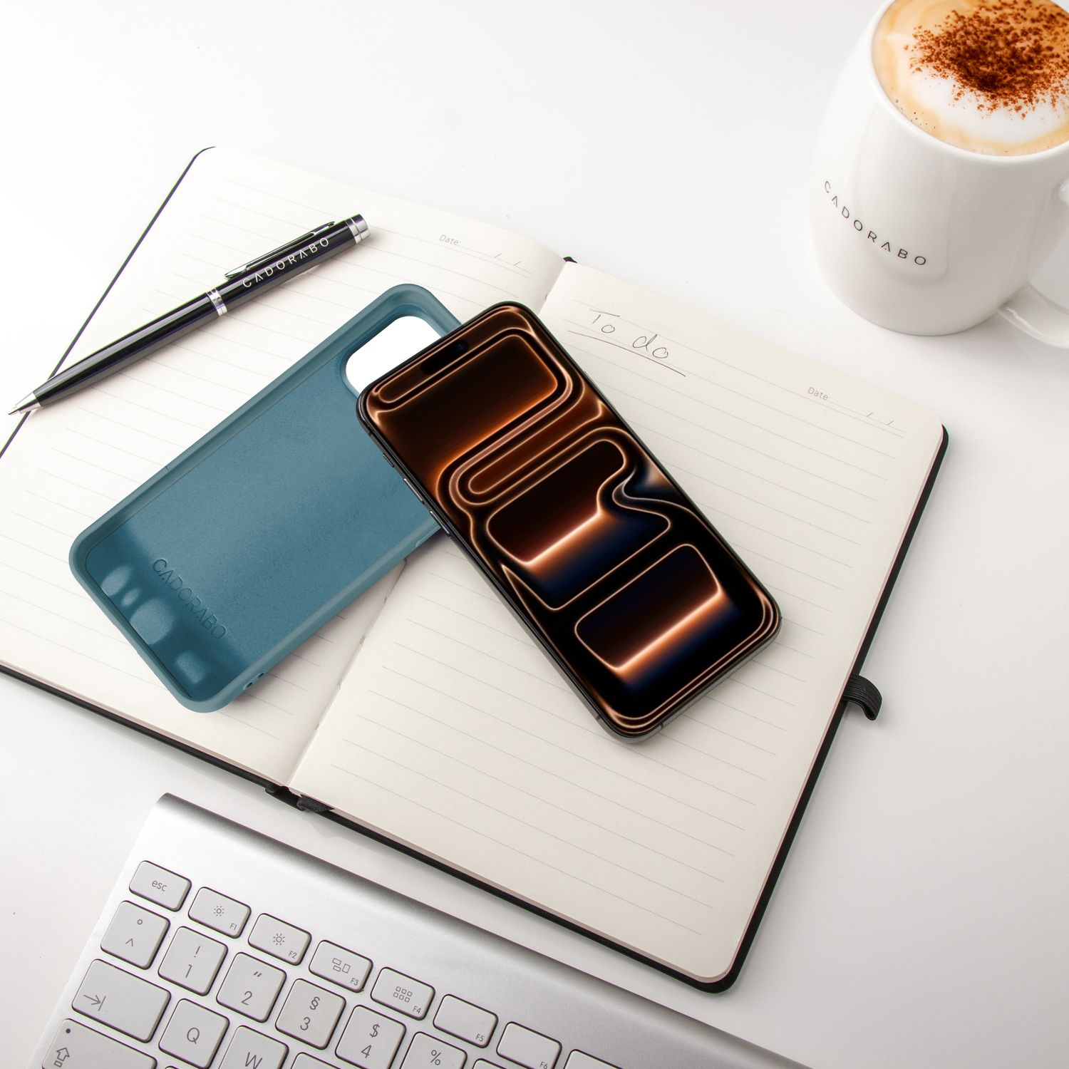 Ein Telefon mit leuchtendem Display liegt auf einem offenen Notizbuch. Ein Stift, eine tealfarbene Handyhülle und eine Kaffeetasse liegen ebenfalls auf dem Schreibtisch.