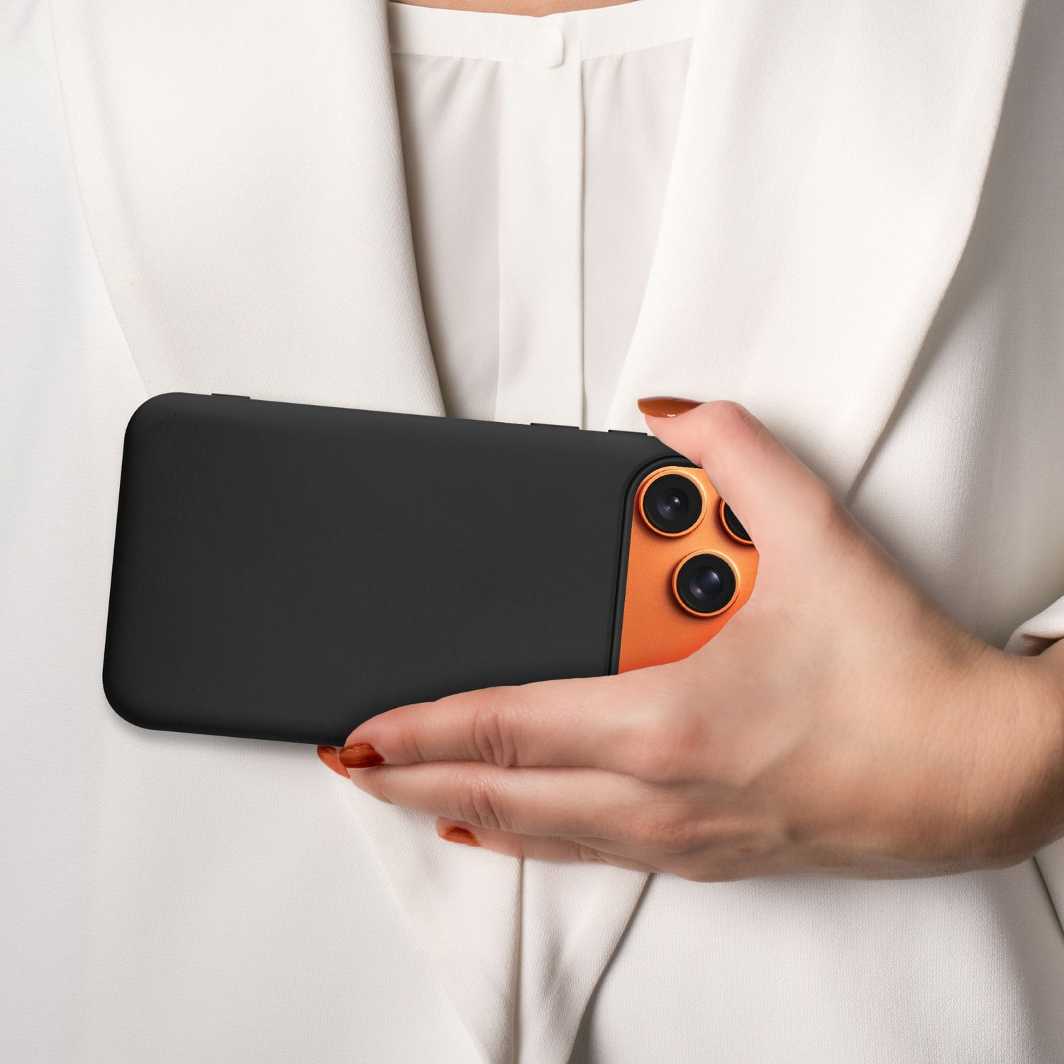 Eine Person hält ein schwarzes Telefon mit orangefarbener Kameraleiste. Die Person trägt einen weißen Blazer, die Hand ist manikürt.