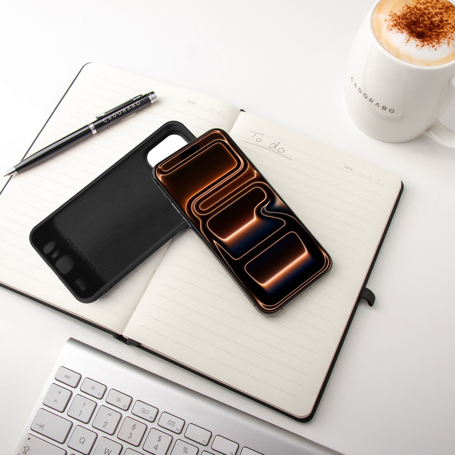 Ein Telefon mit Hülle, Stift, Notizbuch und Kaffeetasse auf einer weißen Oberfläche mit einer Tastatur. Das Telefon zeigt ein beleuchtetes Design.
