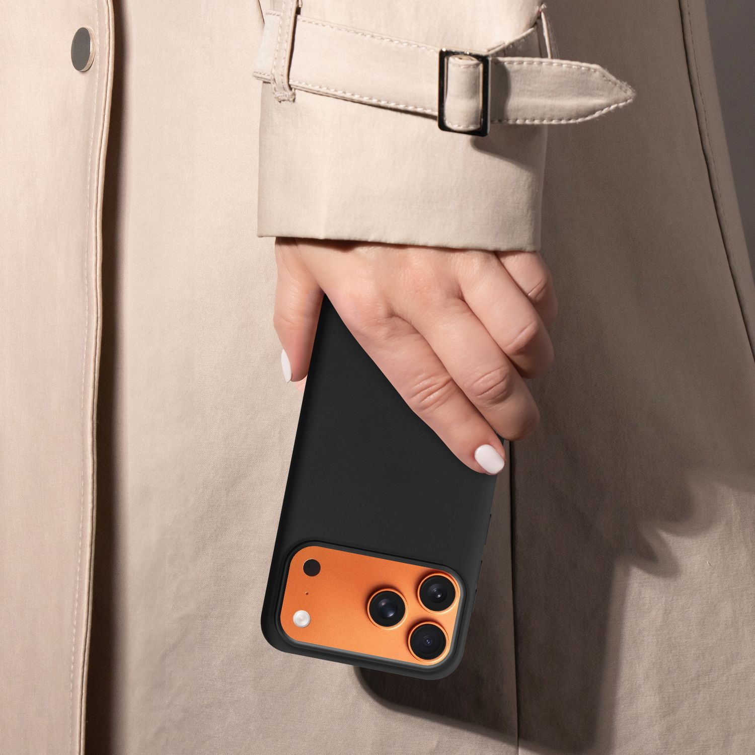 Eine Hand hält ein schwarzes Telefon mit einem orangefarbenen Kameramodul. Die Person trägt einen beigefarbenen Trenchcoat. Der Hintergrund ist neutral.