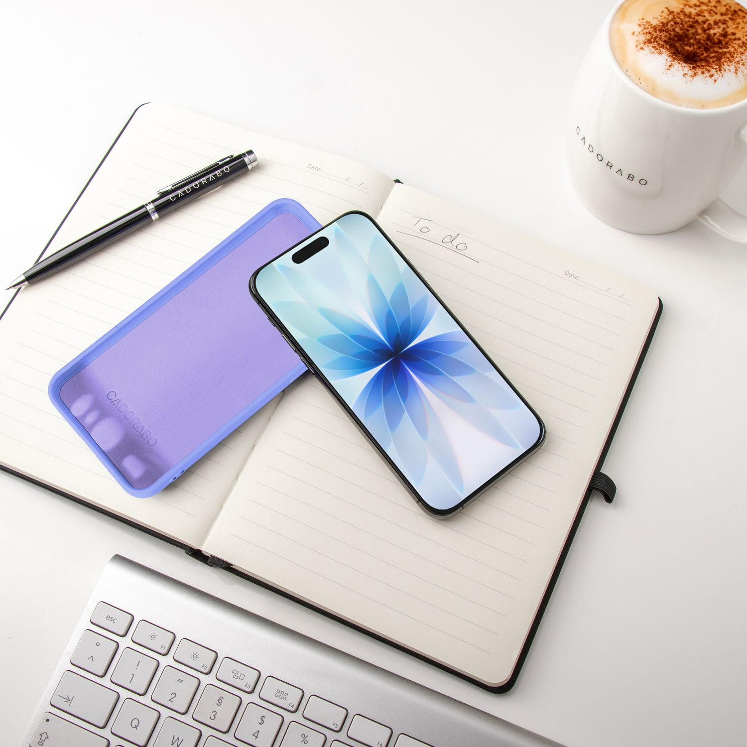Ein Smartphone mit blauem Blumengrund, eine lila Handyhülle, ein Notizblock, ein Stift, eine Tasse Kaffee und eine Tastatur liegen auf weißem Untergrund.