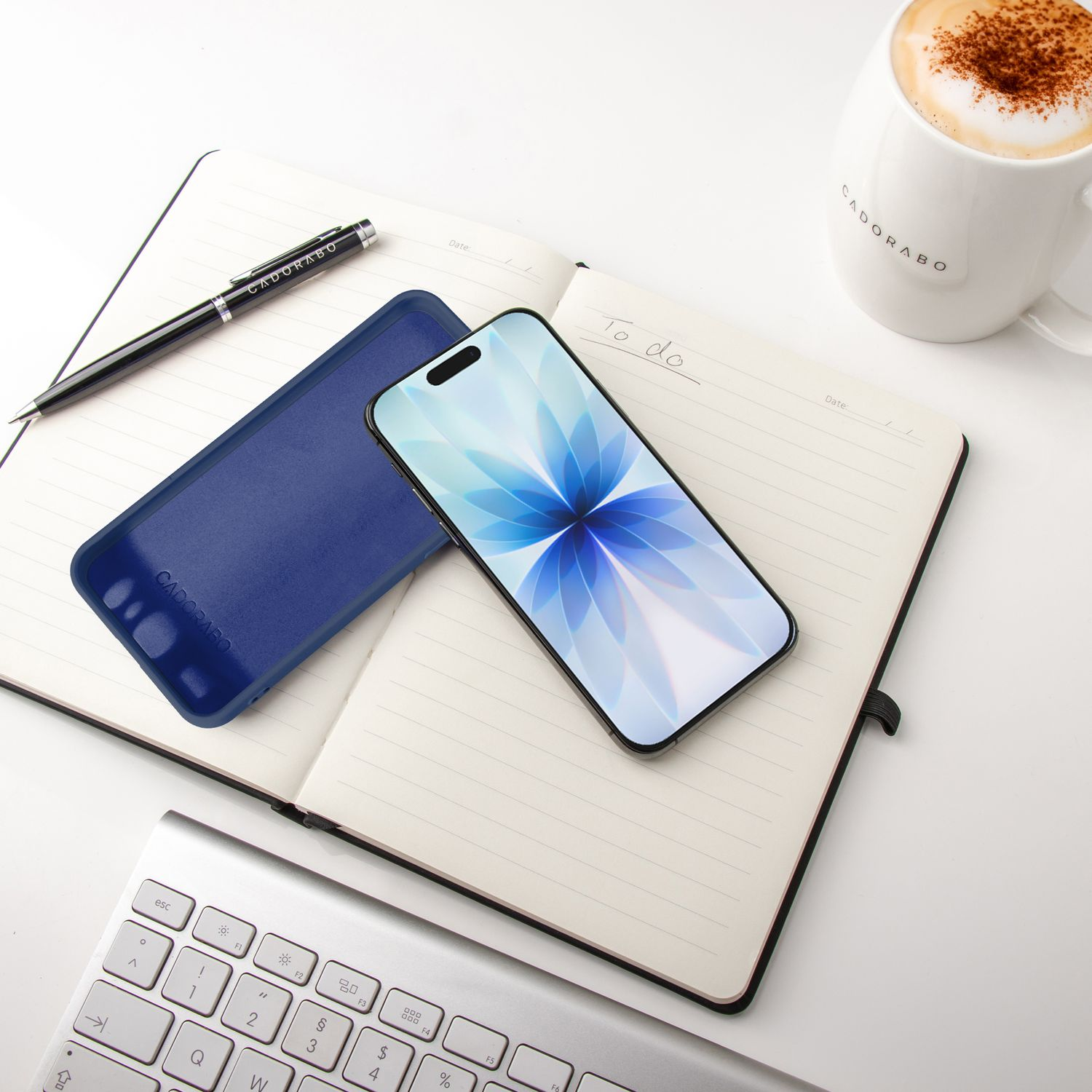 Notizbuch, Telefon, Stift und Kaffeetasse auf weißer Oberfläche. Tastatur im Vordergrund. Telefon zeigt blaues Blumenmuster.