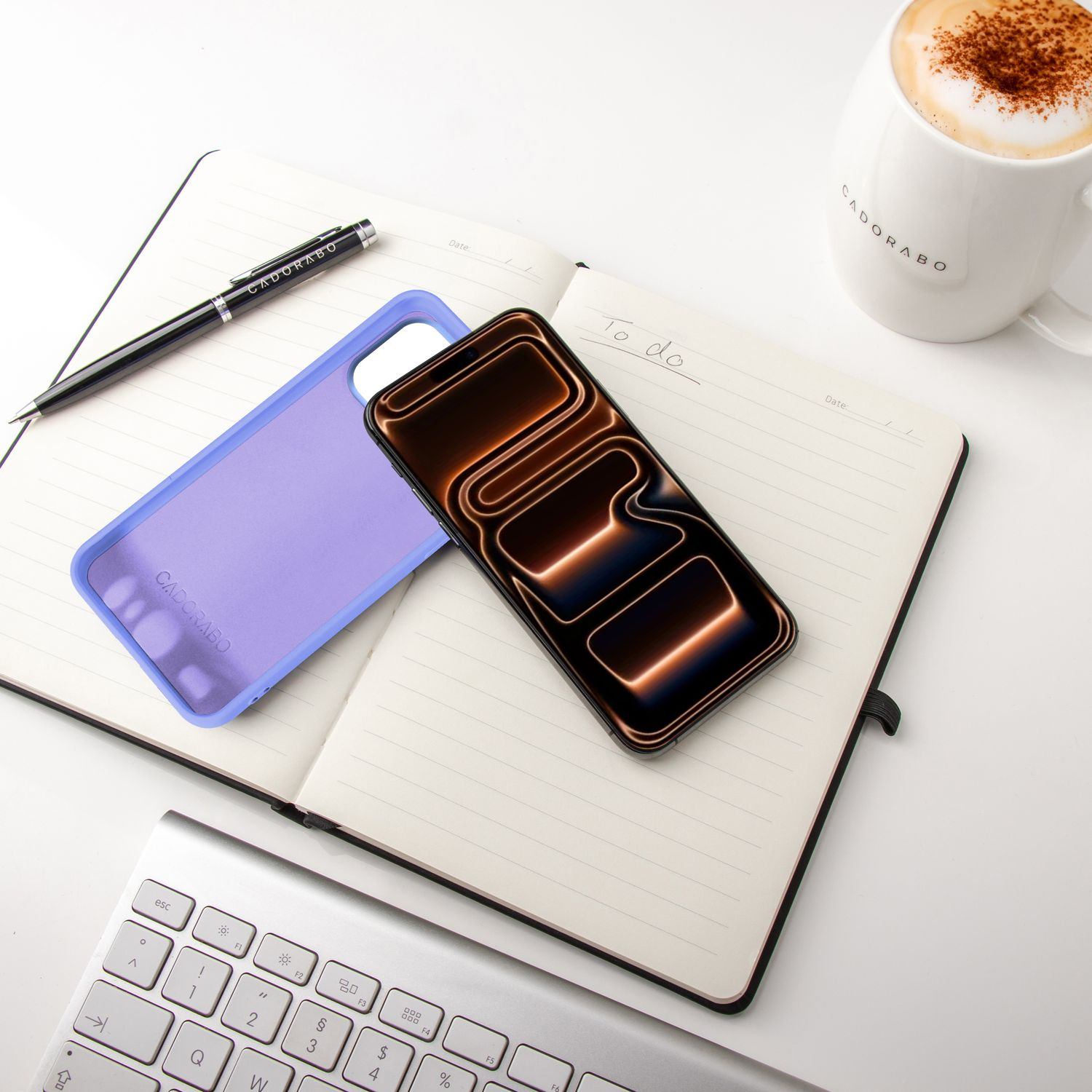 Ein Telefon mit beleuchtetem Bildschirm, Notizbuch, Stift, Kaffeetasse und Tastatur liegen auf einer weißen Oberfläche.