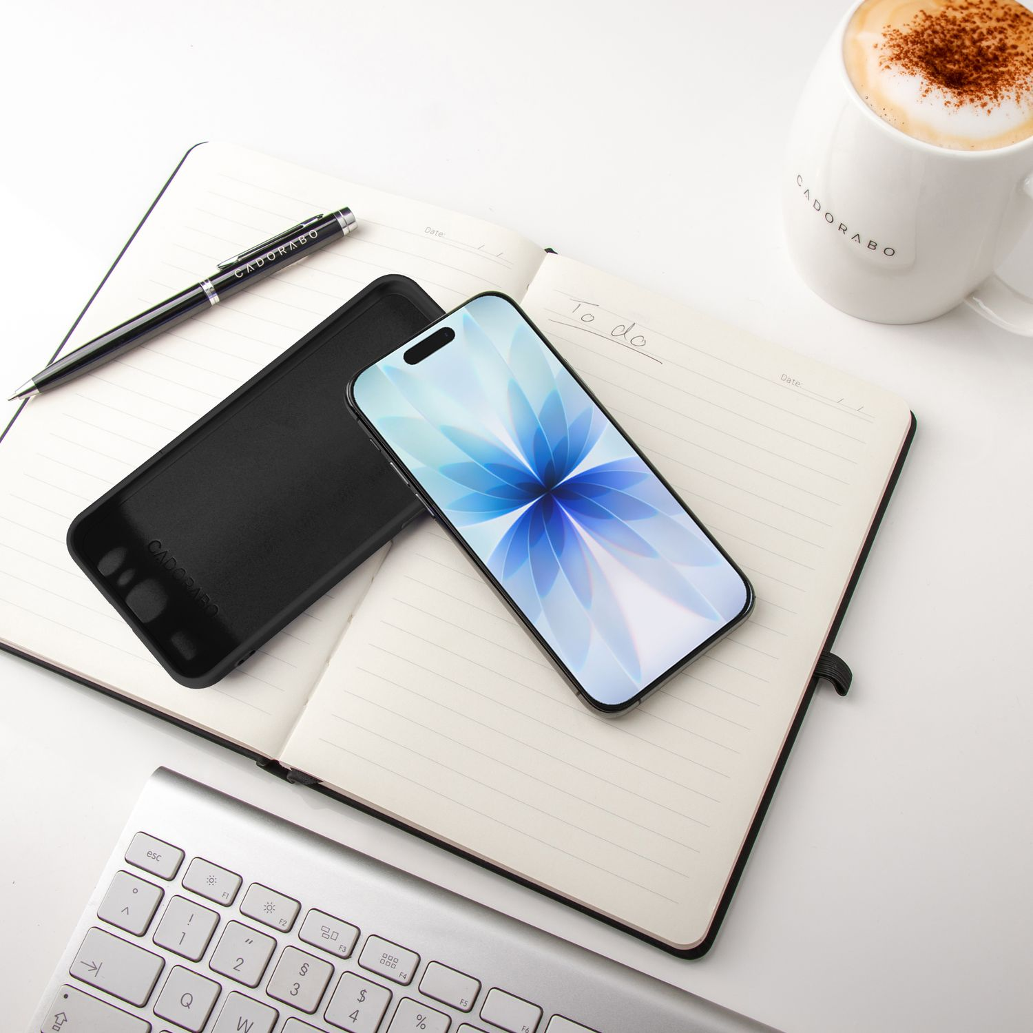 Ein Smartphone mit floralem Hintergrund liegt auf einem offenen Notizbuch. Eine Kaffeetasse, ein Stift und eine Tastatur sind ebenfalls vorhanden.