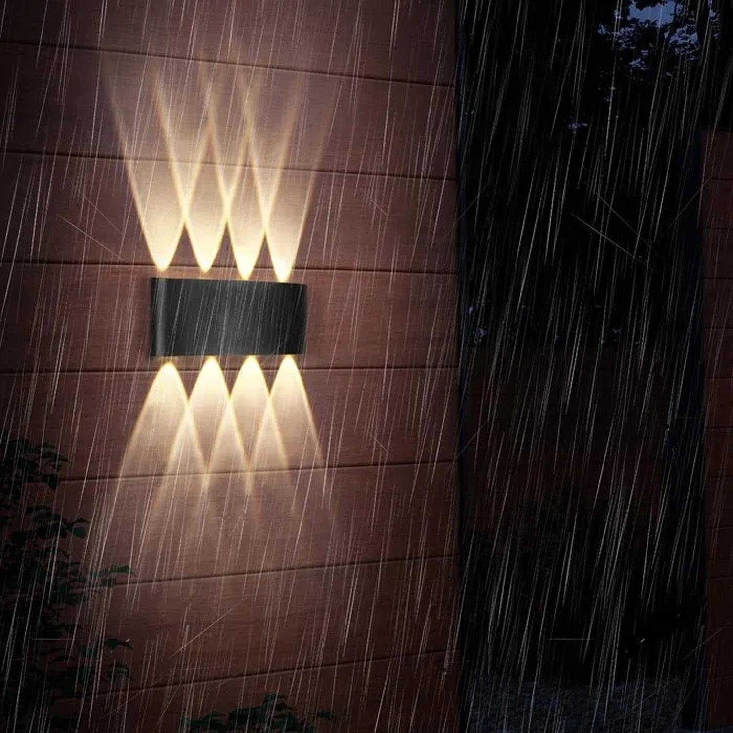 Una lámpara negra montada en la pared ilumina una pared de madera marrón con haces de luz. La lluvia cae por la pared. Planta verde a la izquierda.