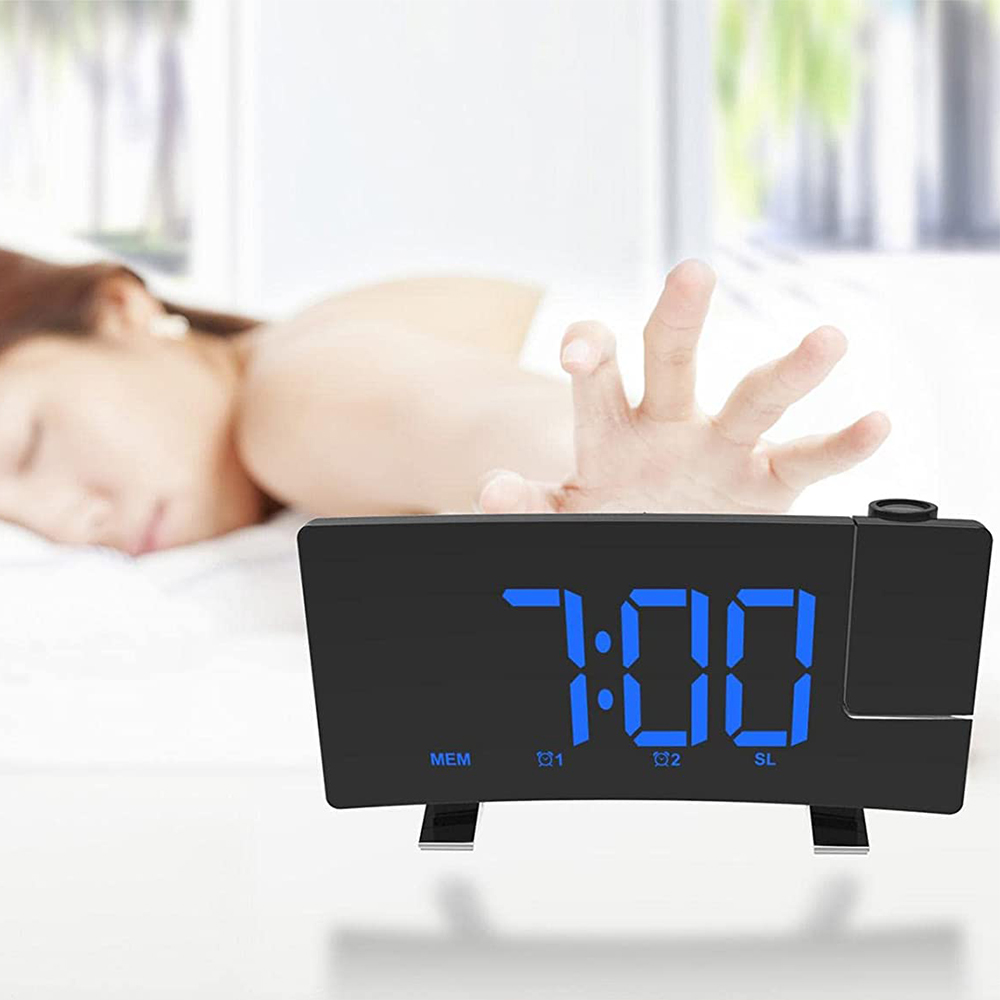 Eine schwarze Digitaluhr zeigt 7:00 Uhr an. Eine Person greift vom Bett zur Uhr. Der Hintergrund zeigt ein Schlafzimmer.